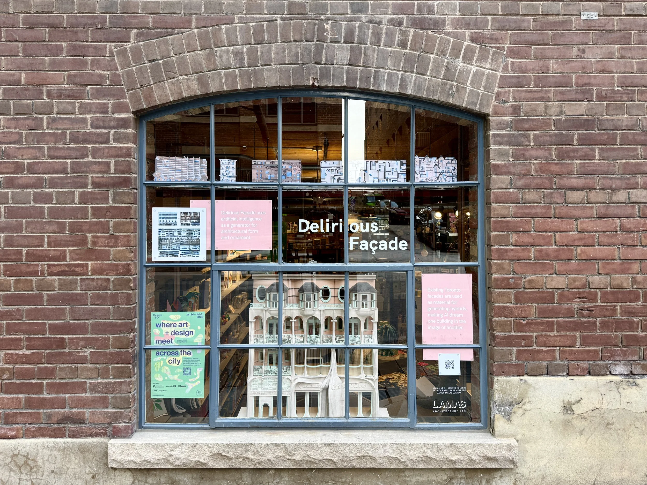 A brick building with a large window displaying architecture models and posters. The window has blue framing and the building has a stone sill. The interior shows displayed models and brochures related to architecture.