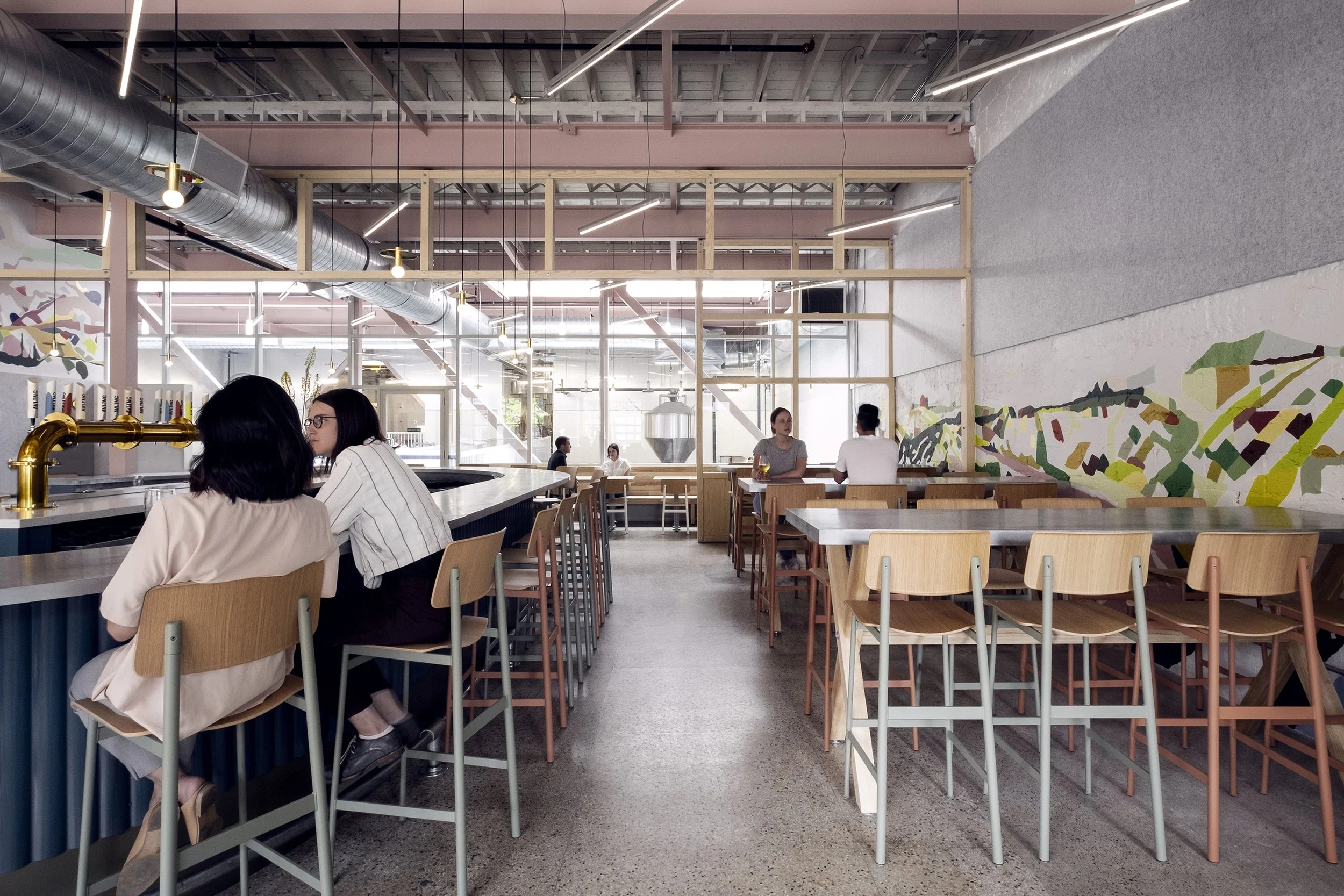 Interior of a modern cafe with wooden chairs and tables, a bar counter with a brass tap, and a colorful mural on the wall. Several people are seated and chatting.
