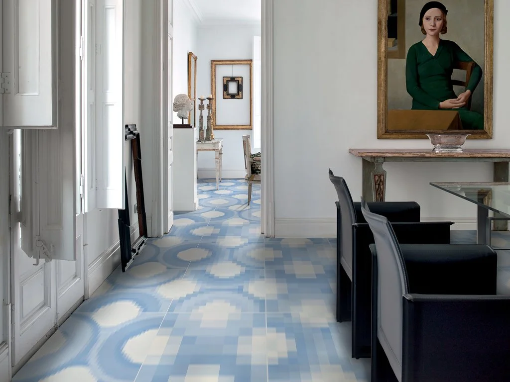 Interior of a stylish home with patterned blue and white floor tiles, white walls, artwork, and furniture including chairs and a glass table.