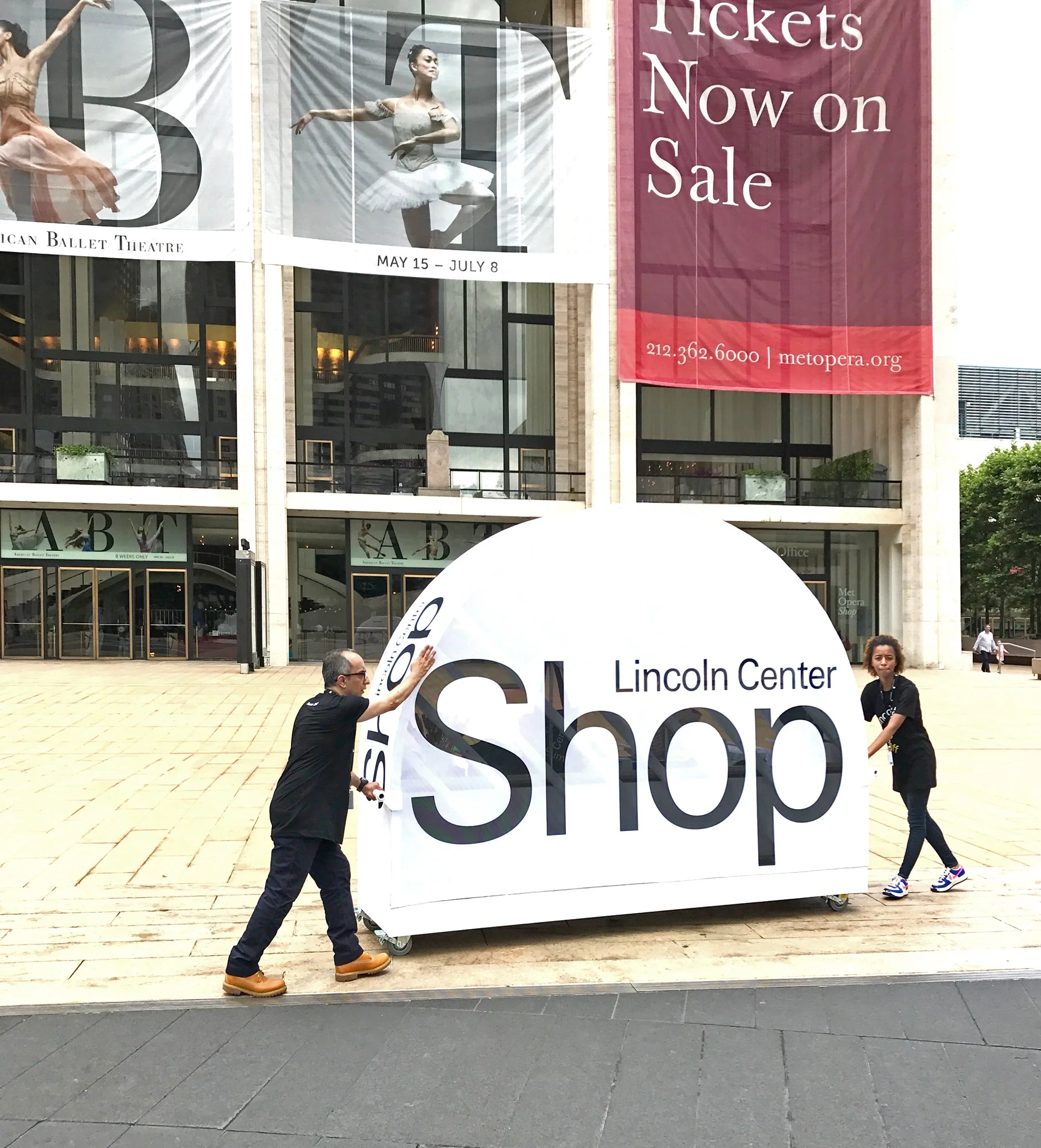 Two people are installing a large white sign that reads 'Lincoln Center Shop' outside a building with advertisements for ballet performances on the windows.
