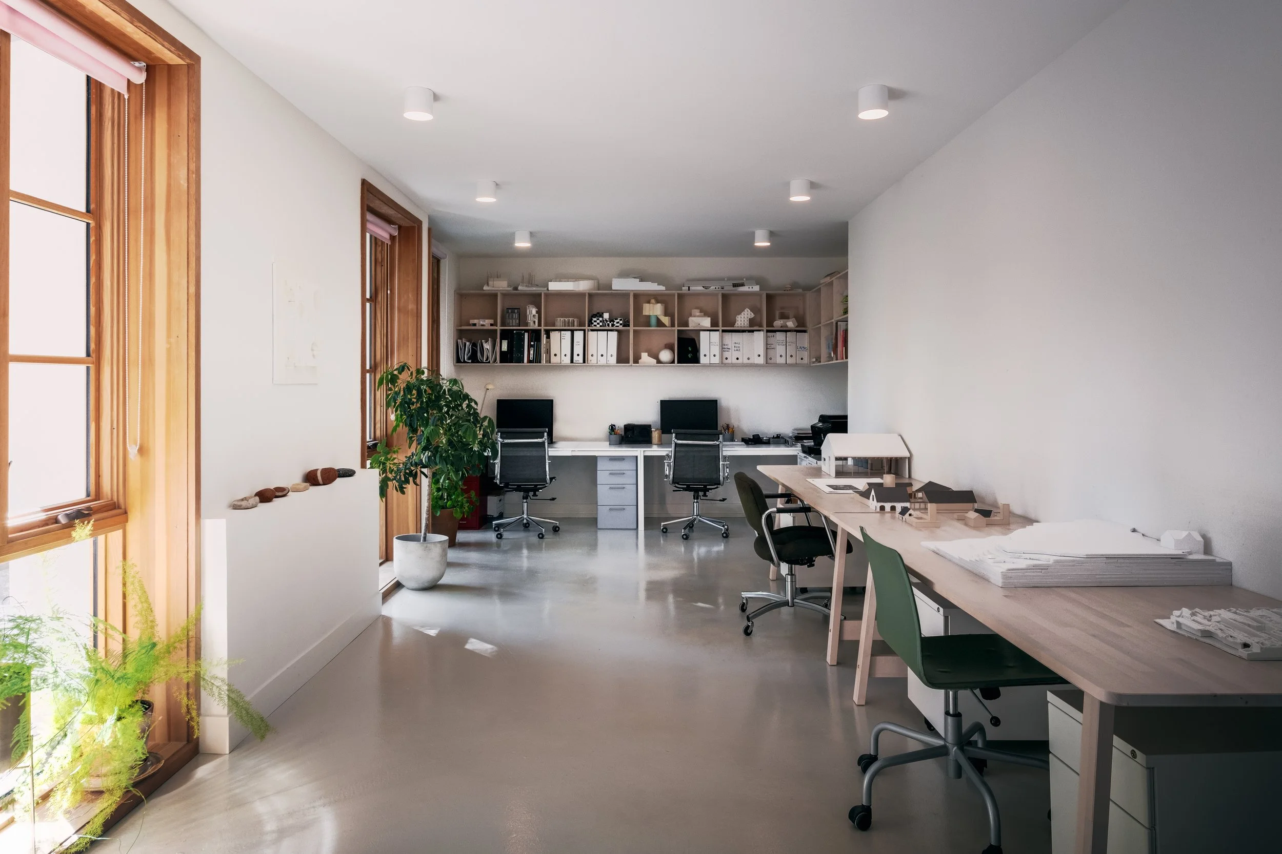 Modern office with wooden framed windows, shelves with binders and office supplies, desks with computers, and chairs in a bright, minimalist design.