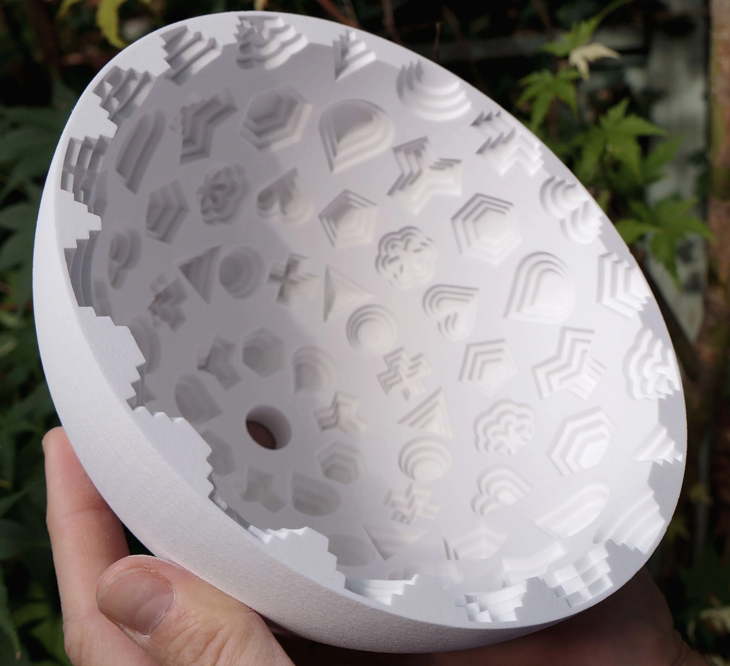 Person holding a round white object with various geometric shapes and patterns cut out, outdoors with green foliage in the background.