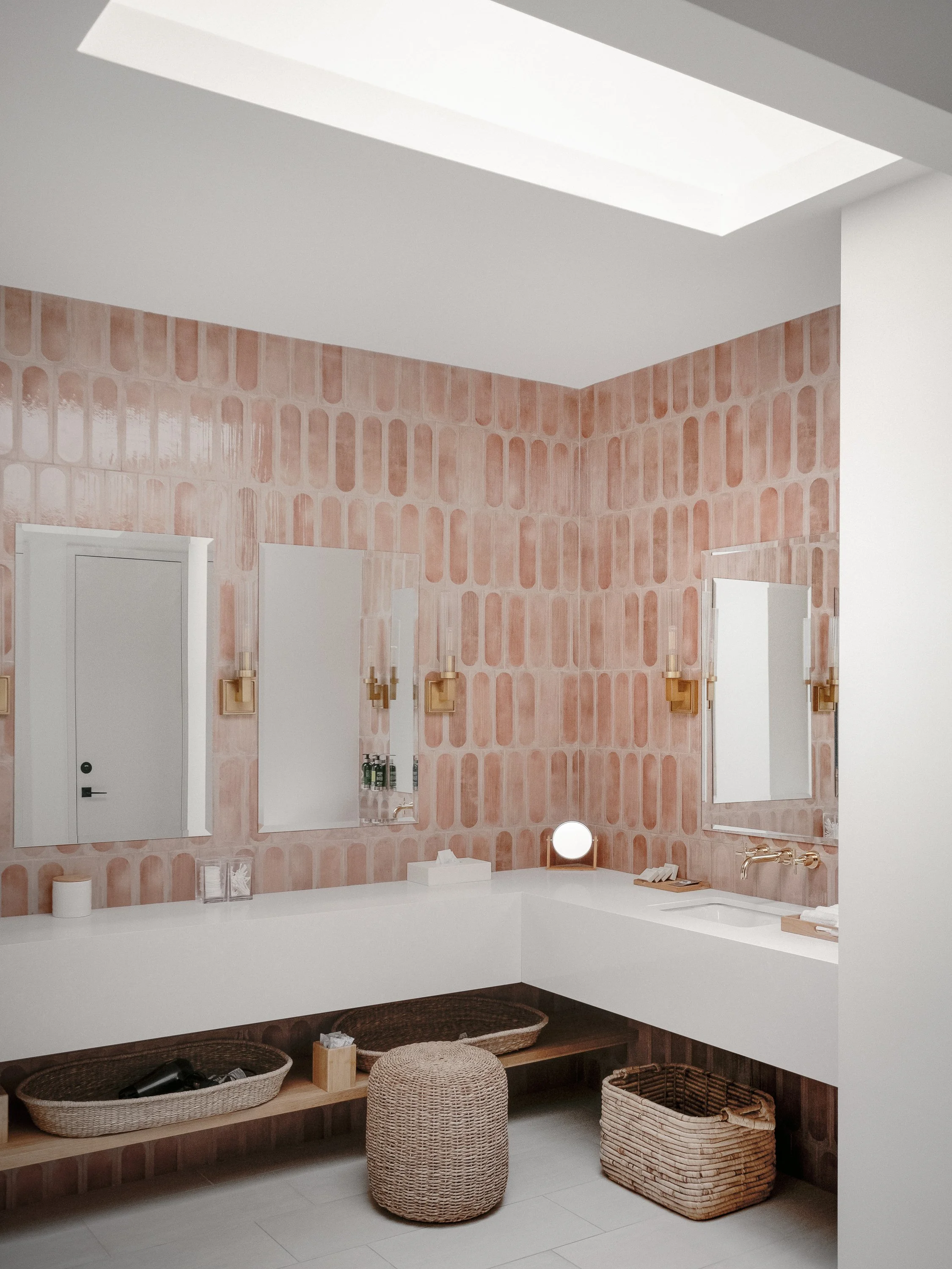 Modern bathroom with pink patterned tiles, white vanity with mirrors, gold light fixtures, and wicker baskets for storage.