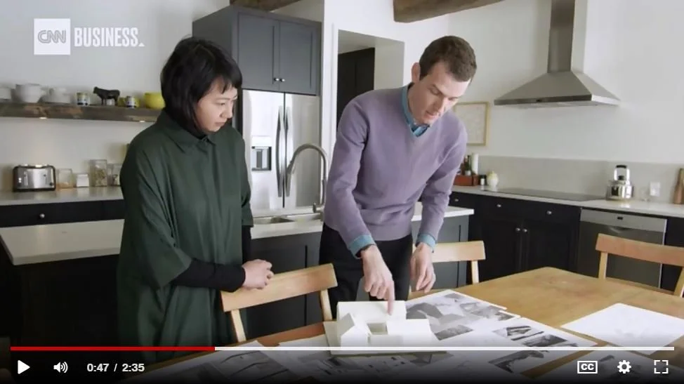 Two people standing around a kitchen table looking at architectural plans or designs.