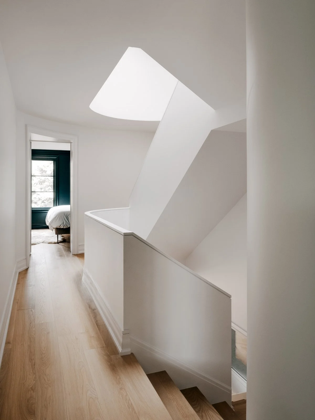 White hallway with staircase leading downstairs, visible bedroom at the end with window and bed.