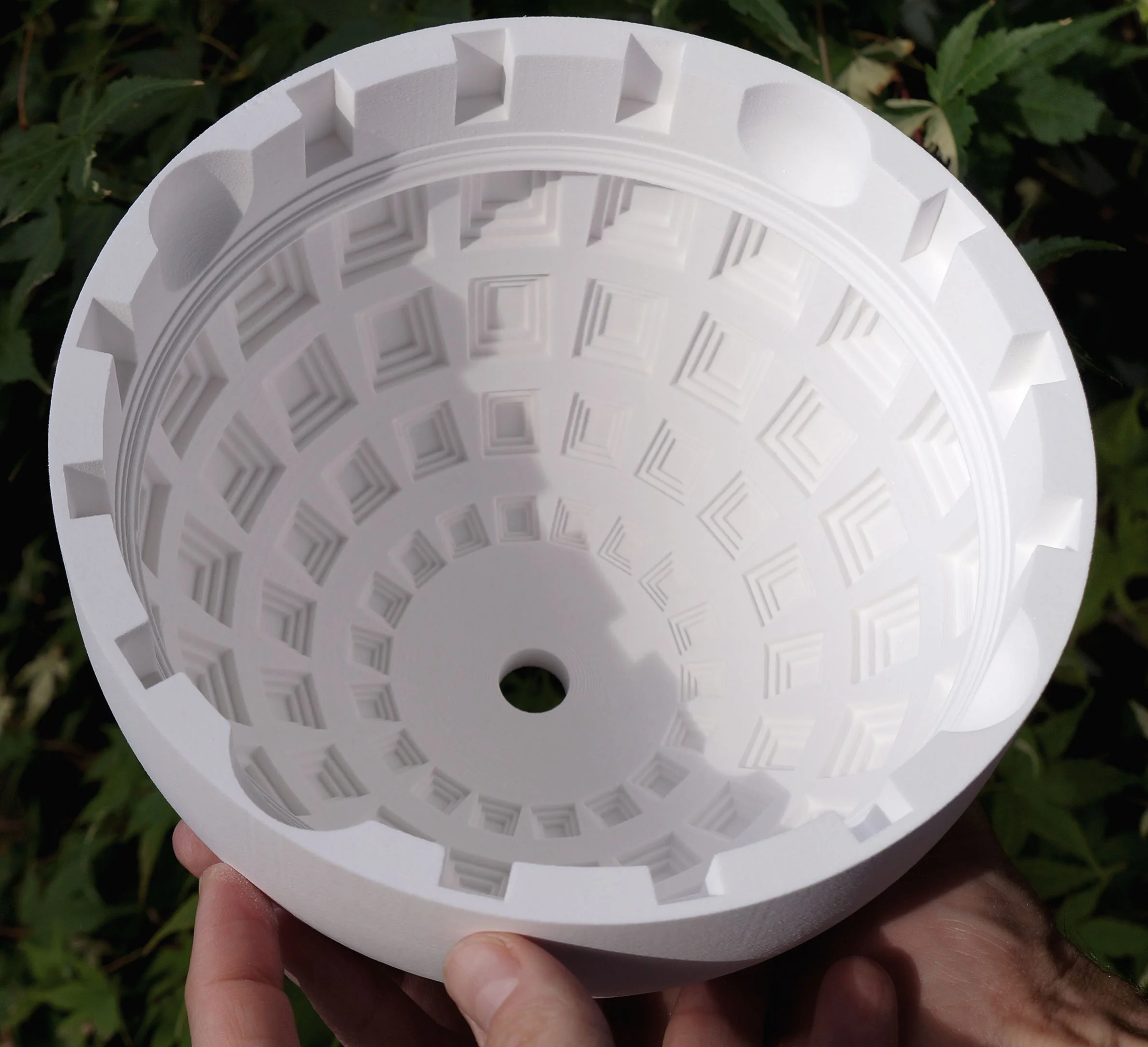 A person holding a white, hollow, round plastic object with a grid-like interior pattern and a small central hole, outdoors with green leaves in the background.