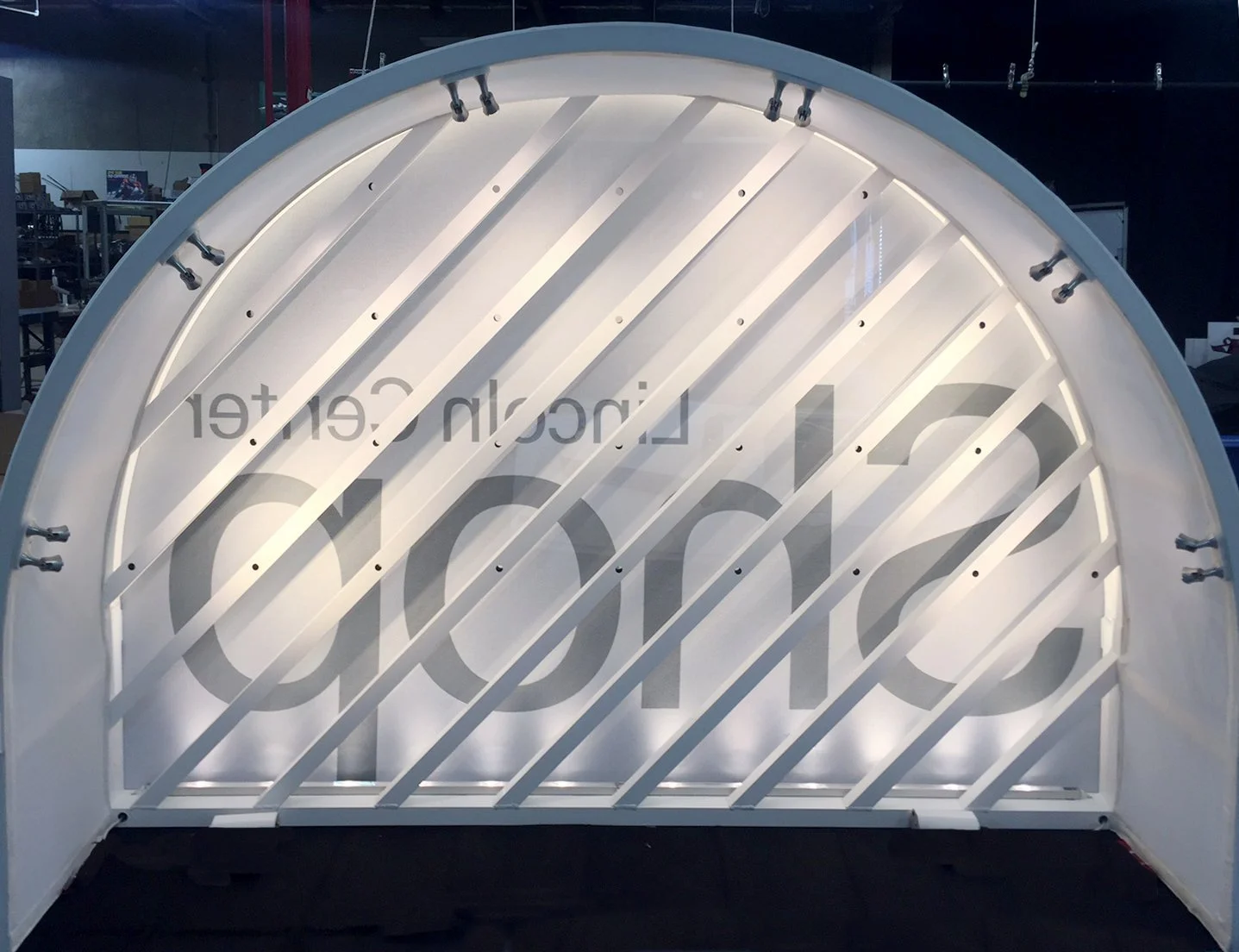 A white, semi-circular, illuminated structure with diagonal slats and faint text behind it.