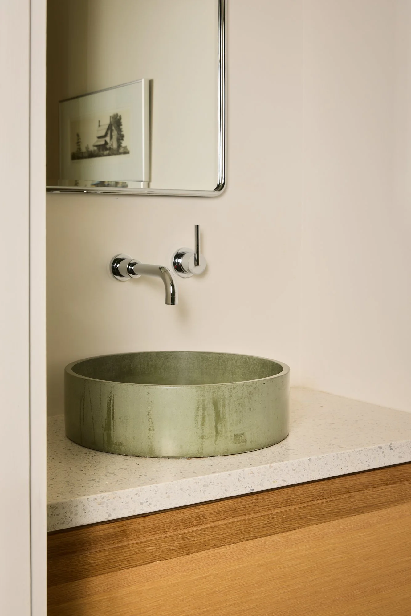 A minimalist bathroom sink with a round, green stone basin, a wall-mounted chrome faucet, a mirror above, and a beige countertop with a wood base.