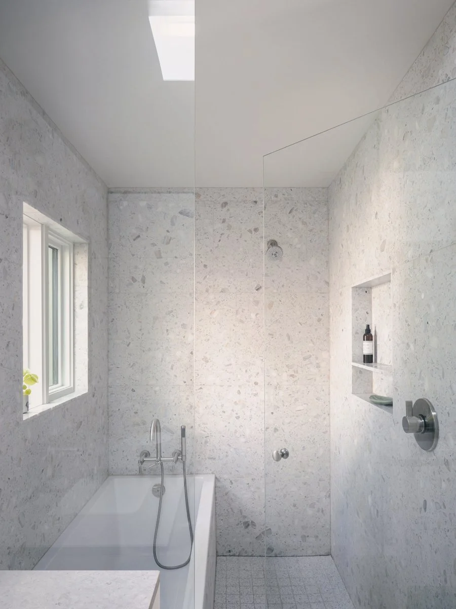 Modern bathroom with white stone walls, a built-in bathtub, a small window, and a separate shower area with a wall niche holding a bottle and a plate, minimalistic design, natural light.