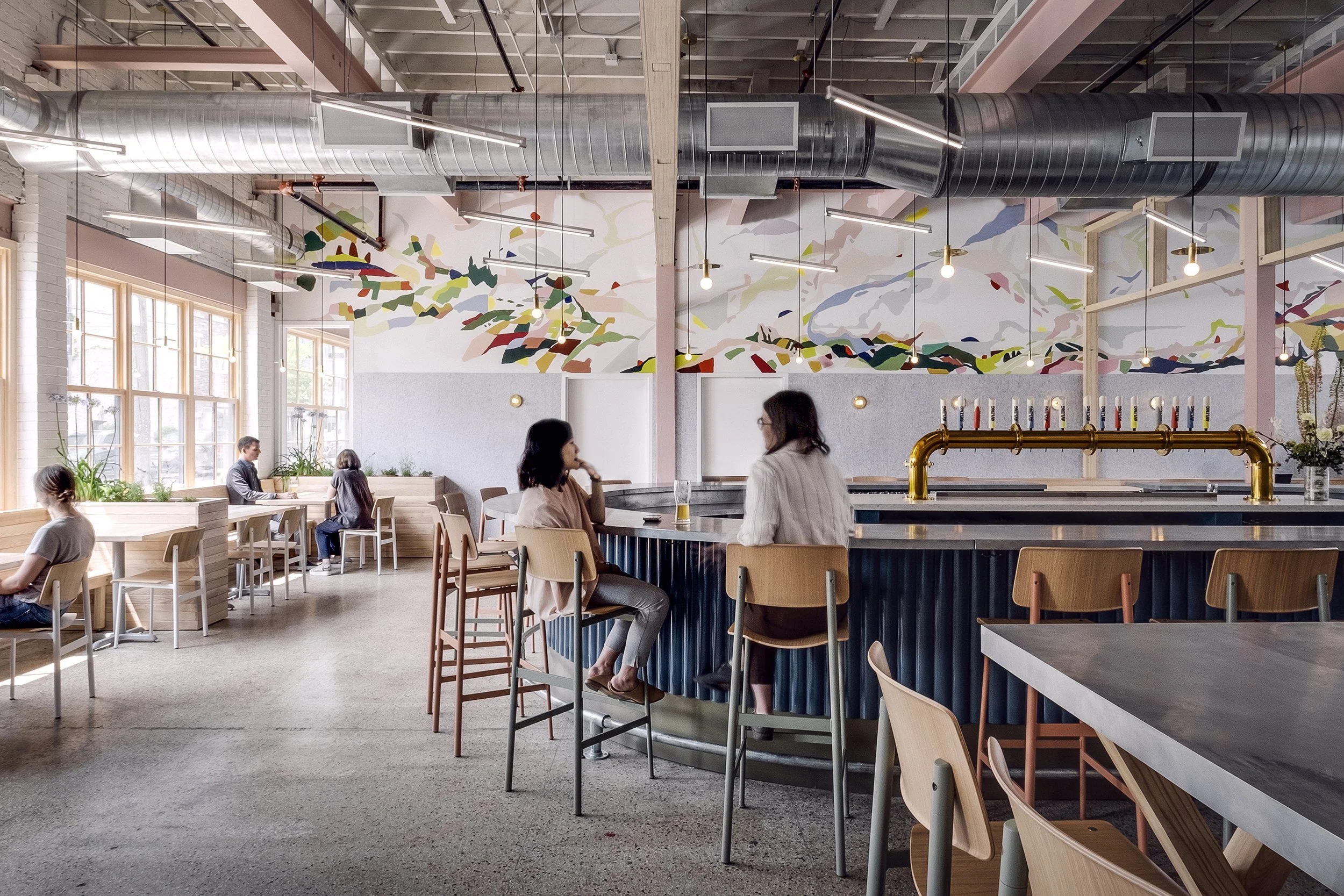 Interior of a modern, brightly lit cafe with large windows, colorful abstract wall art, wooden furniture, and a bar with taps. Several people are seated and conversing.
