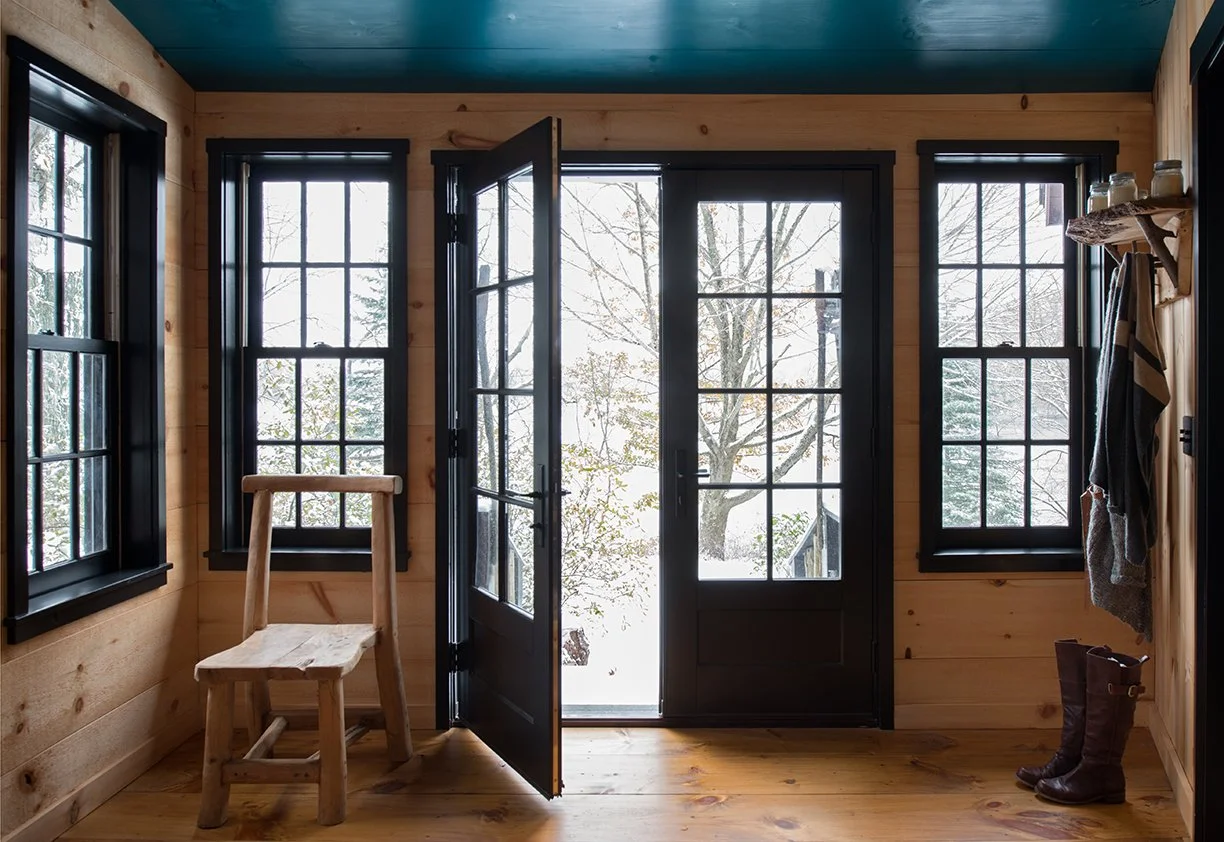 Interior of a cozy wood cabin with black-framed windows and door leading outside to snowy scenery. A small wooden chair is to the left, and a pair of boots and hanging clothes are on the right.