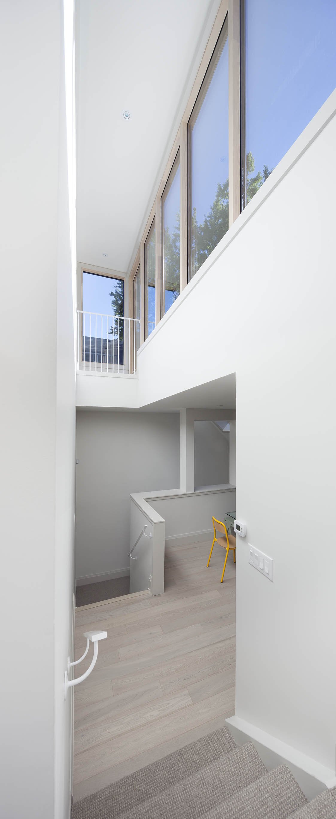 Interior view of a modern, minimalist house with a staircase leading down, large floor-to-ceiling windows, and a yellow chair in a small workspace area.