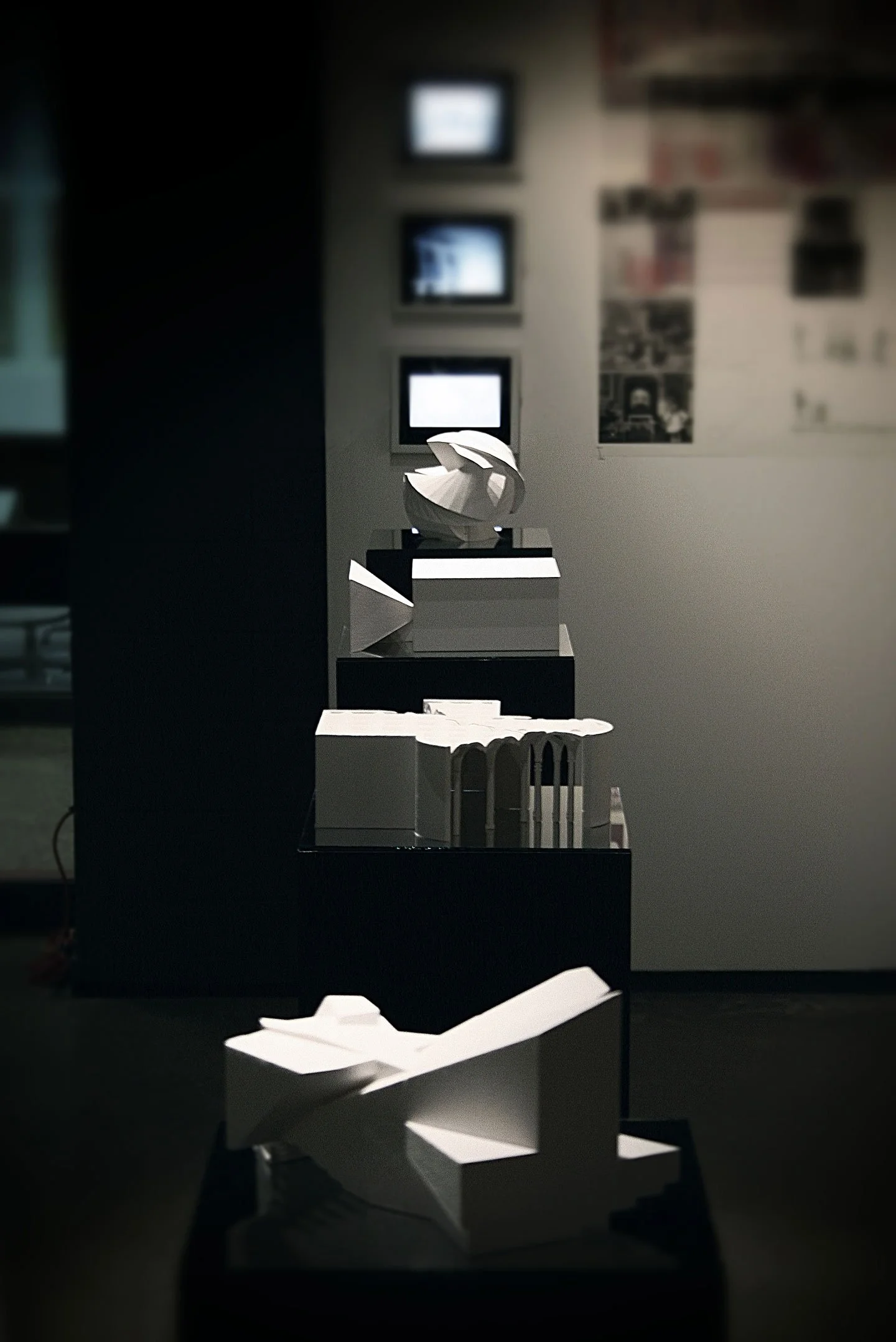 Display of modern architectural models on black pedestals inside a museum or gallery, with framed artwork or informational panels on the wall in the background.