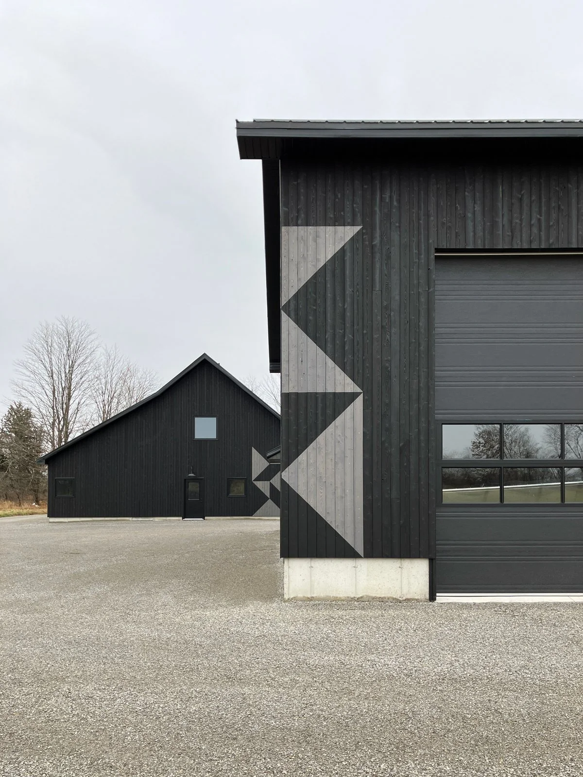 Two black buildings with geometric paint designs on the exterior walls, situated on a gravel lot under an overcast sky.