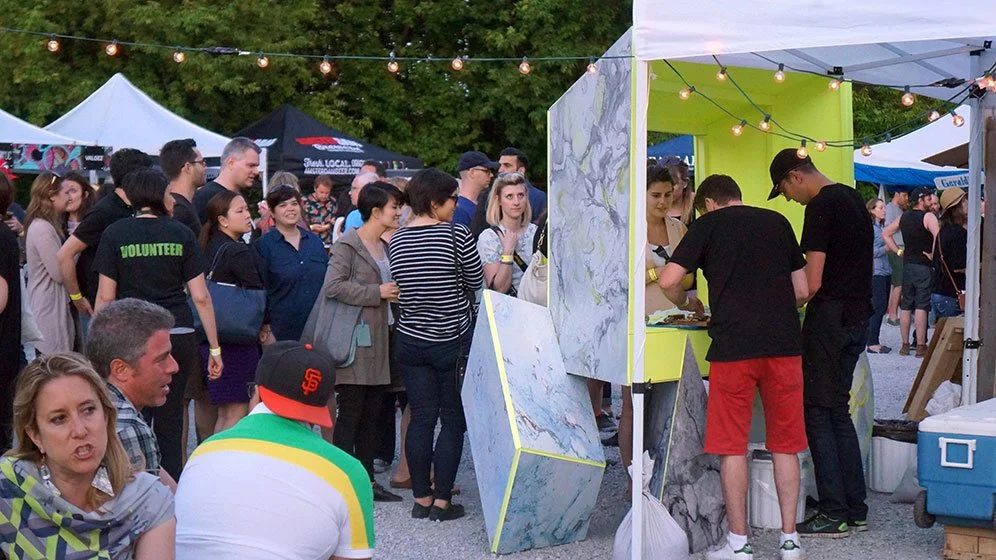 People attending an outdoor event at night, waiting in line at a food or drink stall, with tents and string lights overhead.