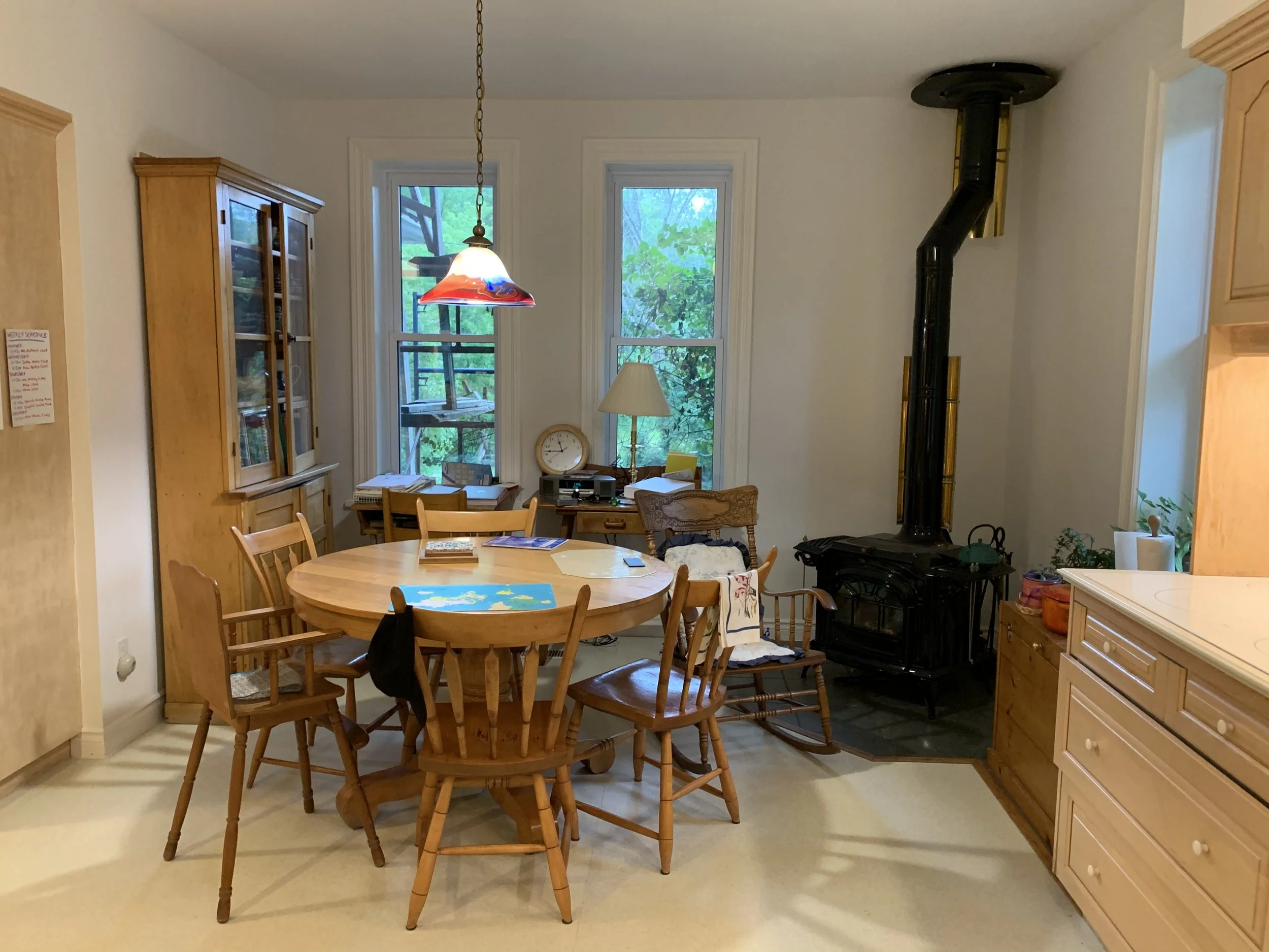 Kitchen with a wooden round dining table surrounded by chairs, two windows showing green foliage outside, and a black wood stove with a tall pipe. The room has wooden cabinets and a bookcase, with a hanging light fixture above the table.