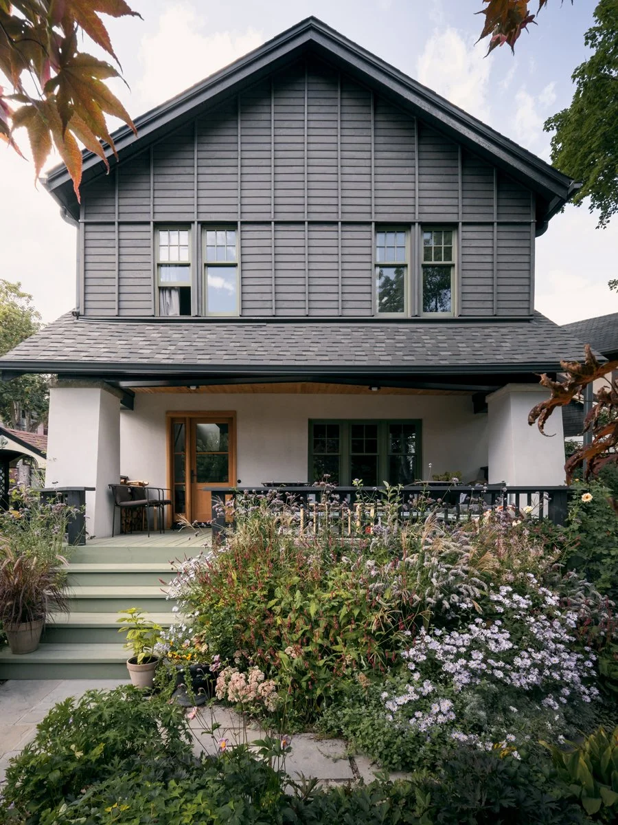 Front view of a two-story house with a dark-gray roof and siding, white walls on the lower level, a wooden door, and a front porch with plants and flowers in pots and beds.