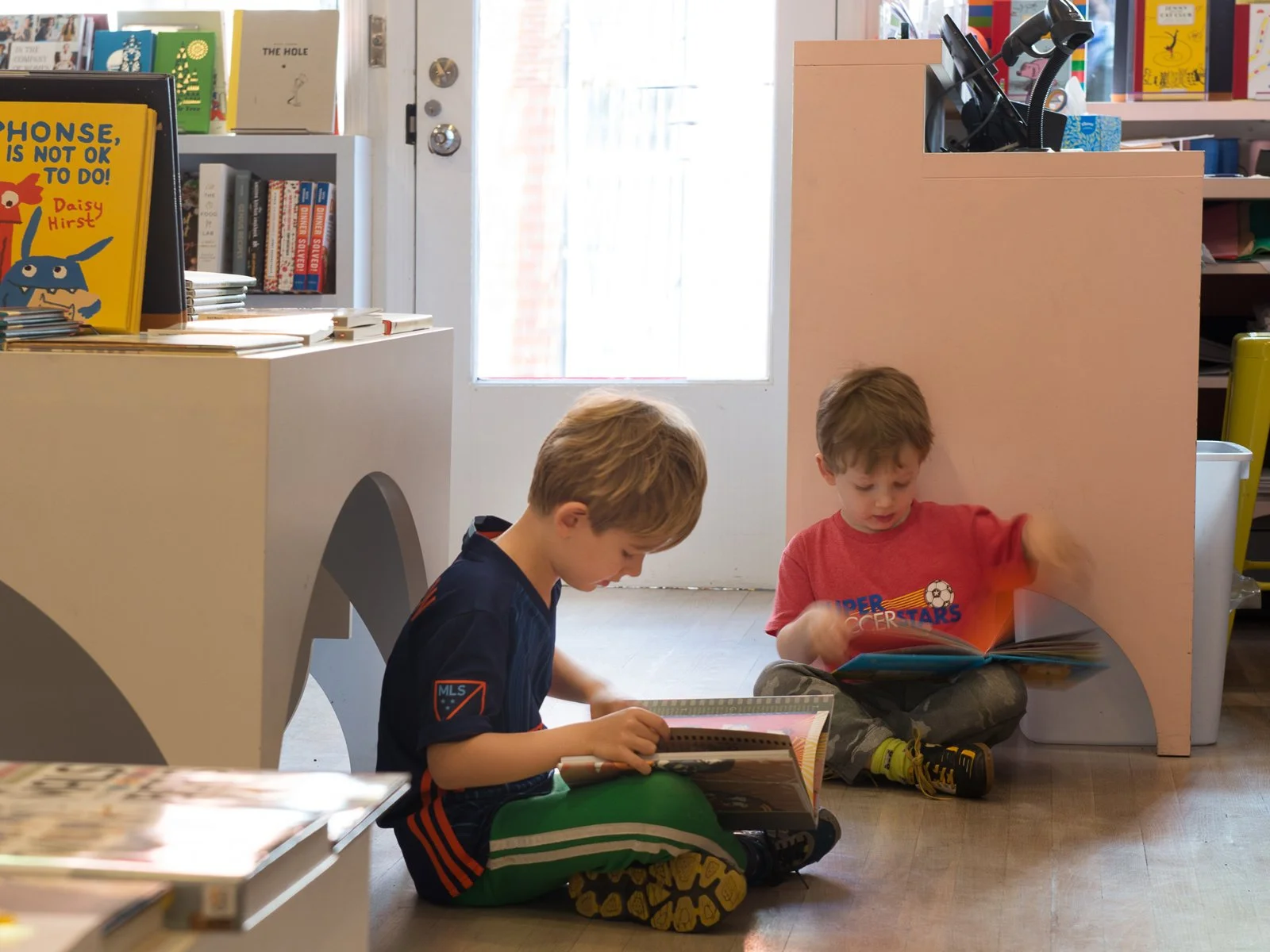 Two young boys sit on the floor in a bookstore, each reading a book. One boy wears a black and green sports jersey and leggings, and the other wears a red t-shirt with a soccer graphic. Behind them, bookshelves are filled with colorful books, and there is a pink partition and door with sunlight coming through.