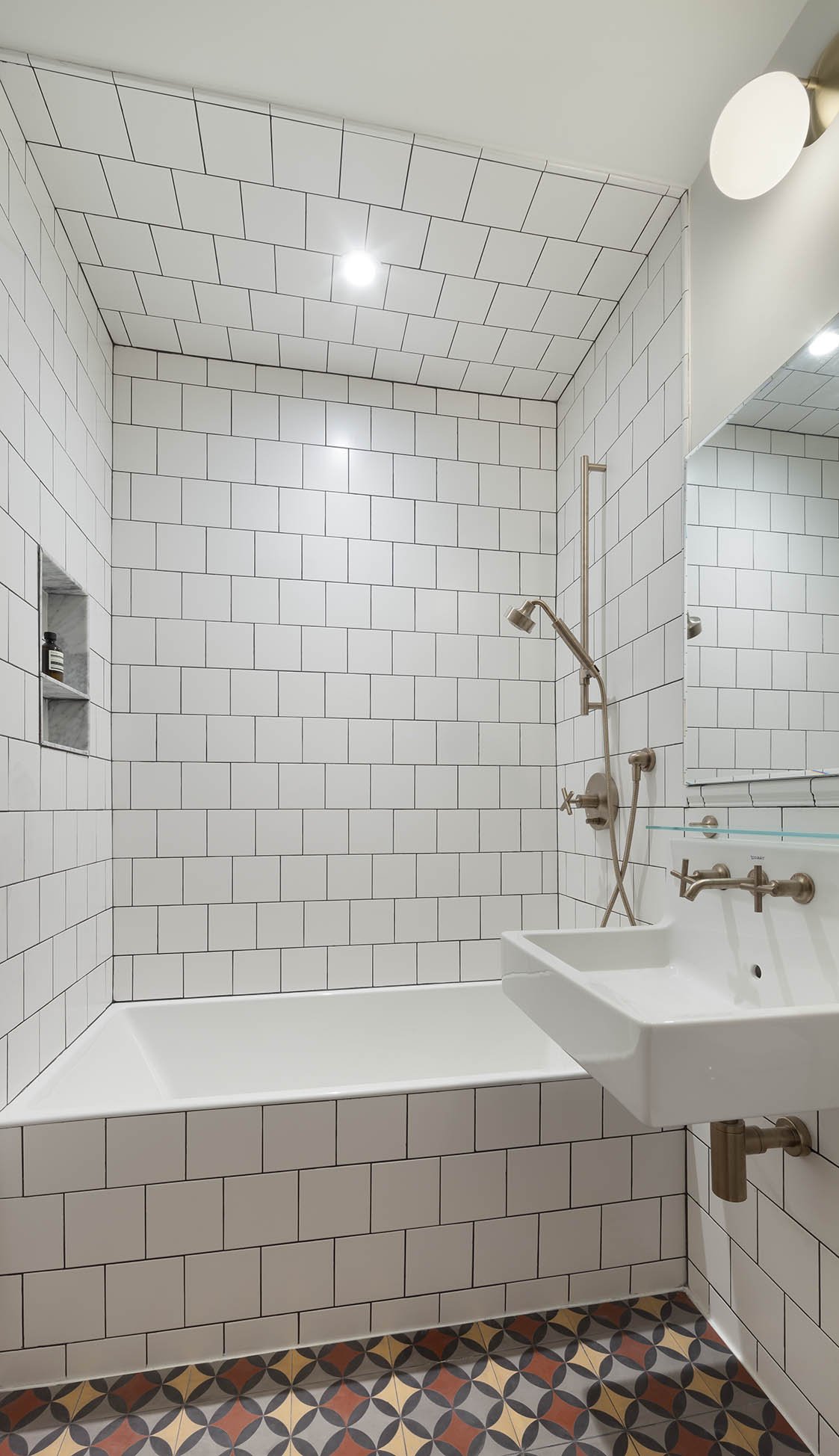 A modern bathroom with white subway tile walls and ceiling, a small bathtub, a wall-mounted sink with a brass faucet, a large mirror, and patterned tile flooring.