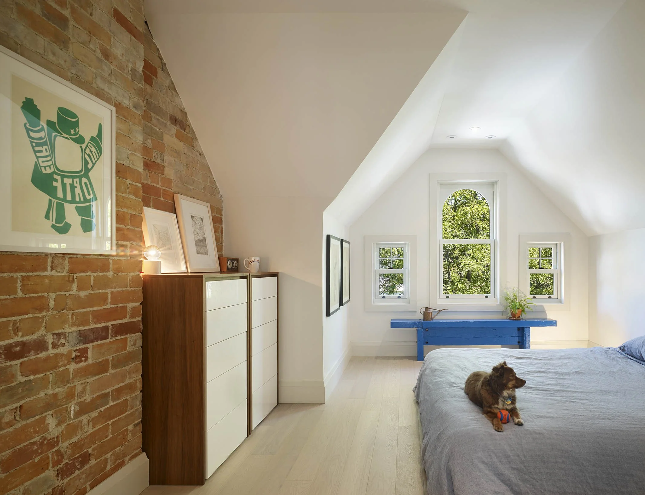 Bright attic bedroom with large window, white walls, exposed brick wall, blue bench, dog with ball on bed, and art and plants decor.