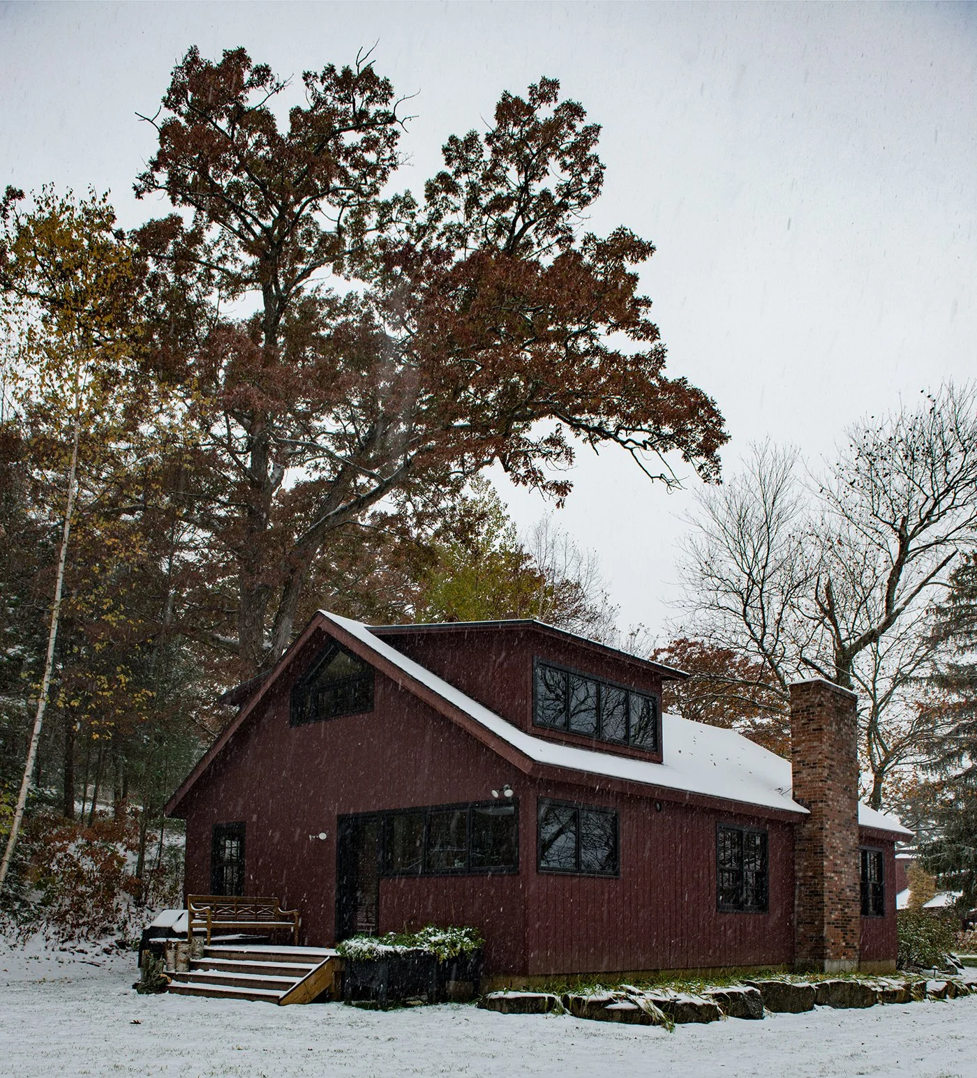 A red house with black window frames and a brick chimney, surrounded by snow and tall trees in a wintery landscape.