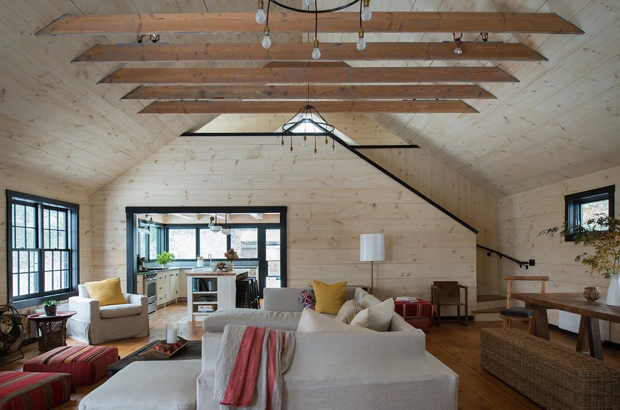 Living room with beige walls and sloped ceiling, featuring large windows, white sofas, colorful pillows, wooden floor, modern lighting fixtures, and a view into a kitchen area.