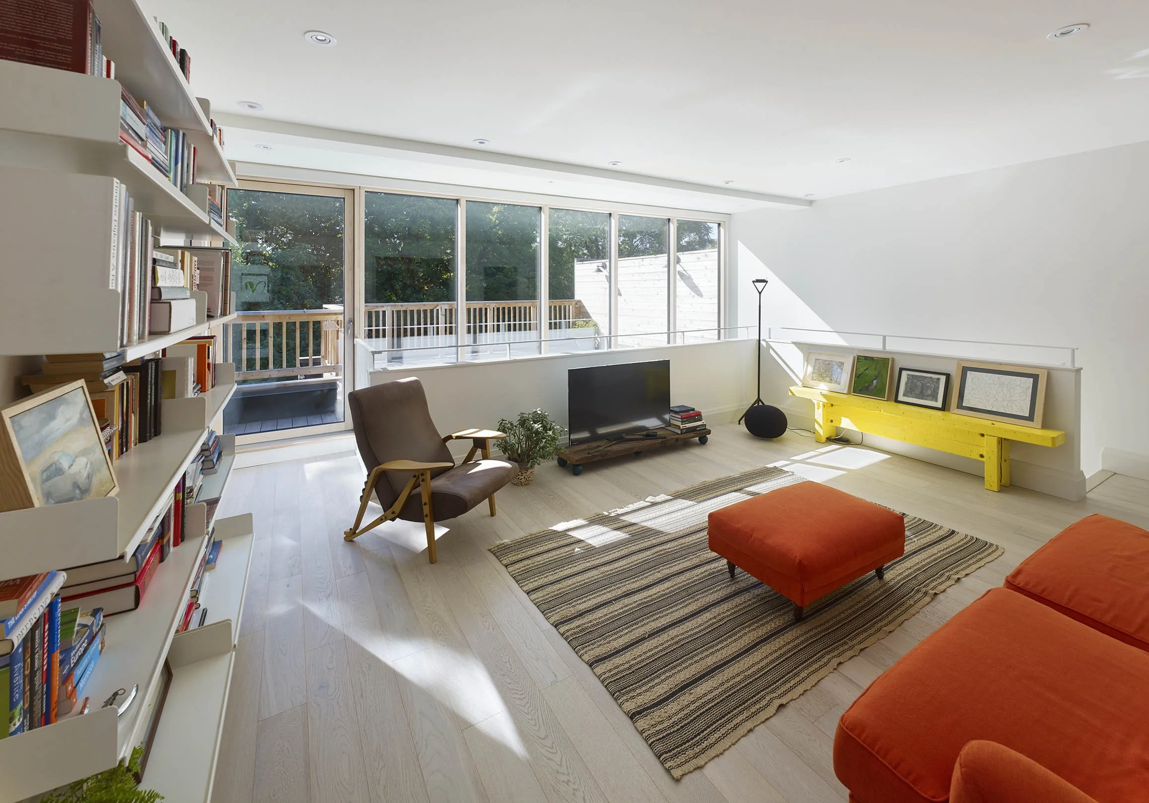 Bright living room with large windows, white walls, a brown armchair, a black TV on a wooden stand, a yellow bench with framed pictures, an orange ottoman, an orange sofa, a striped rug, a tall black floor lamp, and a sliding glass door leading to a balcony with trees outside.