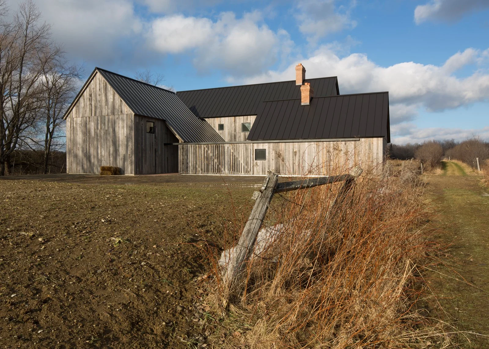 A rustic barn with weathered wooden walls and a black metal roof, situated on a rural landscape with a dirt path and blue sky with scattered clouds.