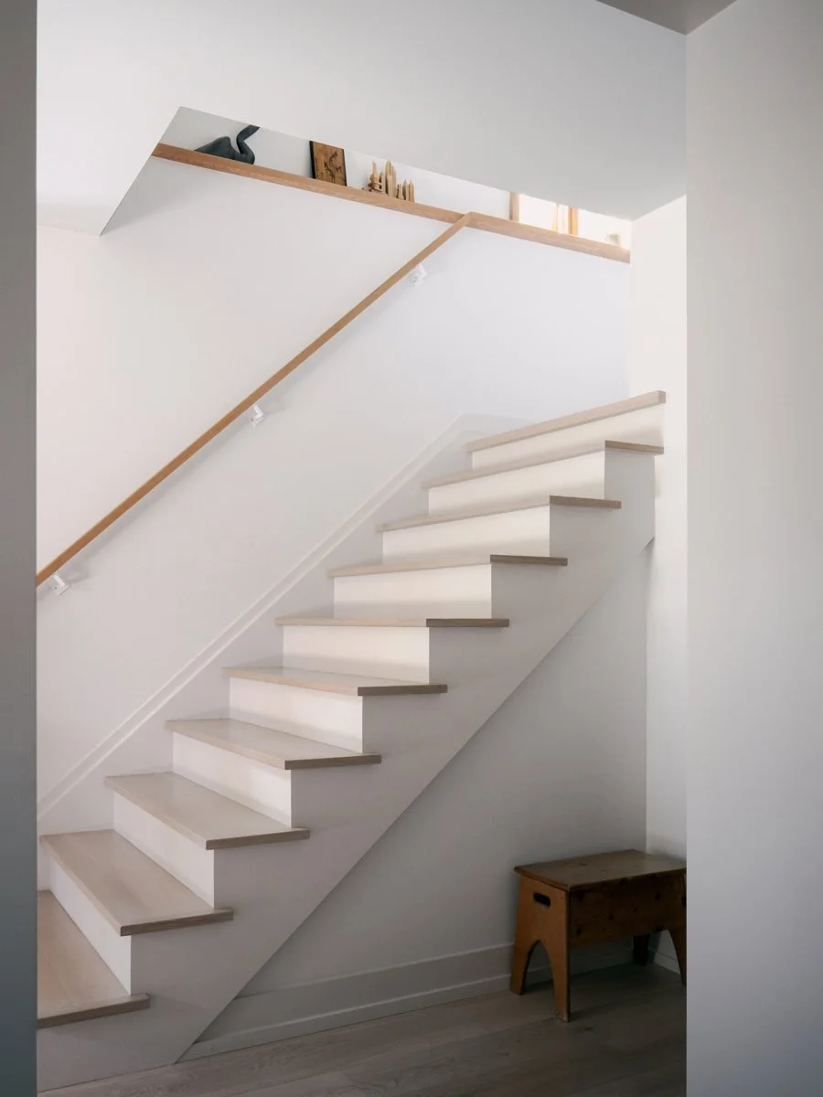 Minimalist staircase with white risers and light wood treads, a simple wooden handrail, and a small wooden bench beneath the stairs in a modern white interior.