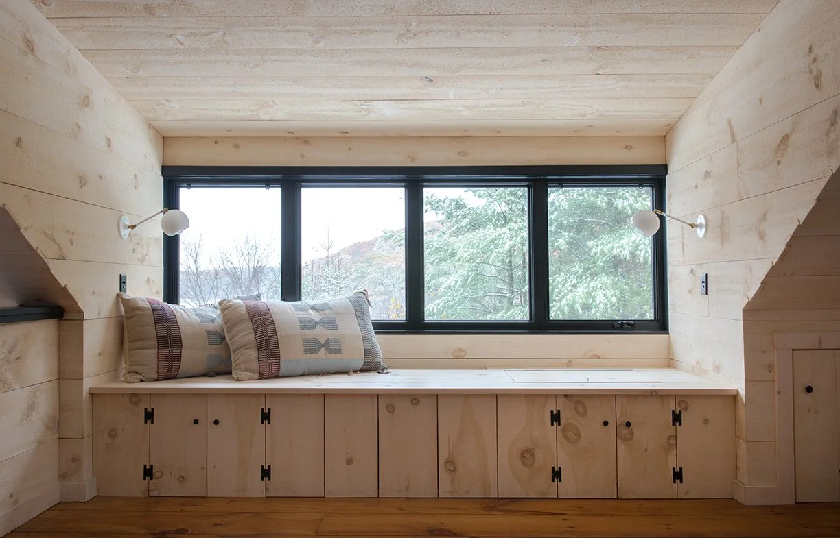 Wood-paneled window seat with cushions, built into a room with wood walls and ceiling, overlooking a view of trees outside.