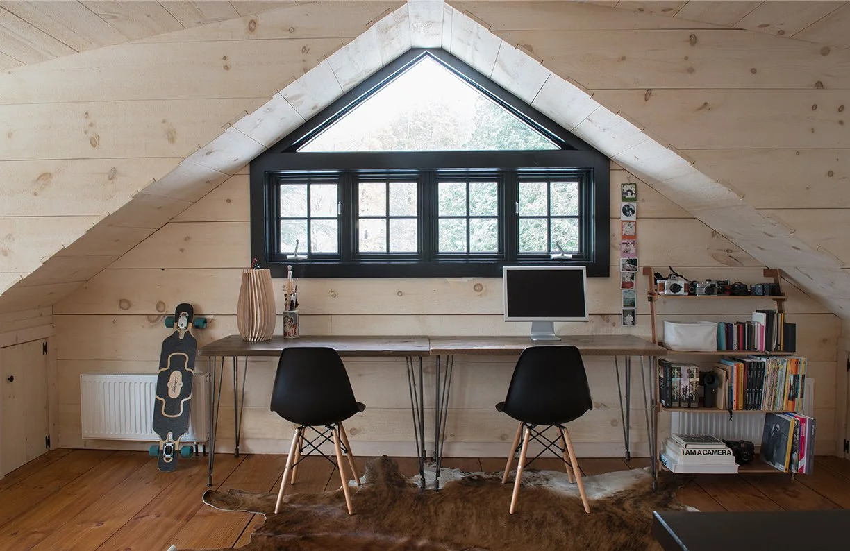 Home office setup with a sloped ceiling, a large triangular window, and a black framed window underneath. There is a long wooden desk with two black chairs, one with a computer monitor. To the left, a skateboard leans against a white radiator, and next to it, a tall wooden vase with art supplies. On the right, a wooden bookshelf holds cameras, books, and decorative items, including a sign that says 'I AM A CAMERA'.