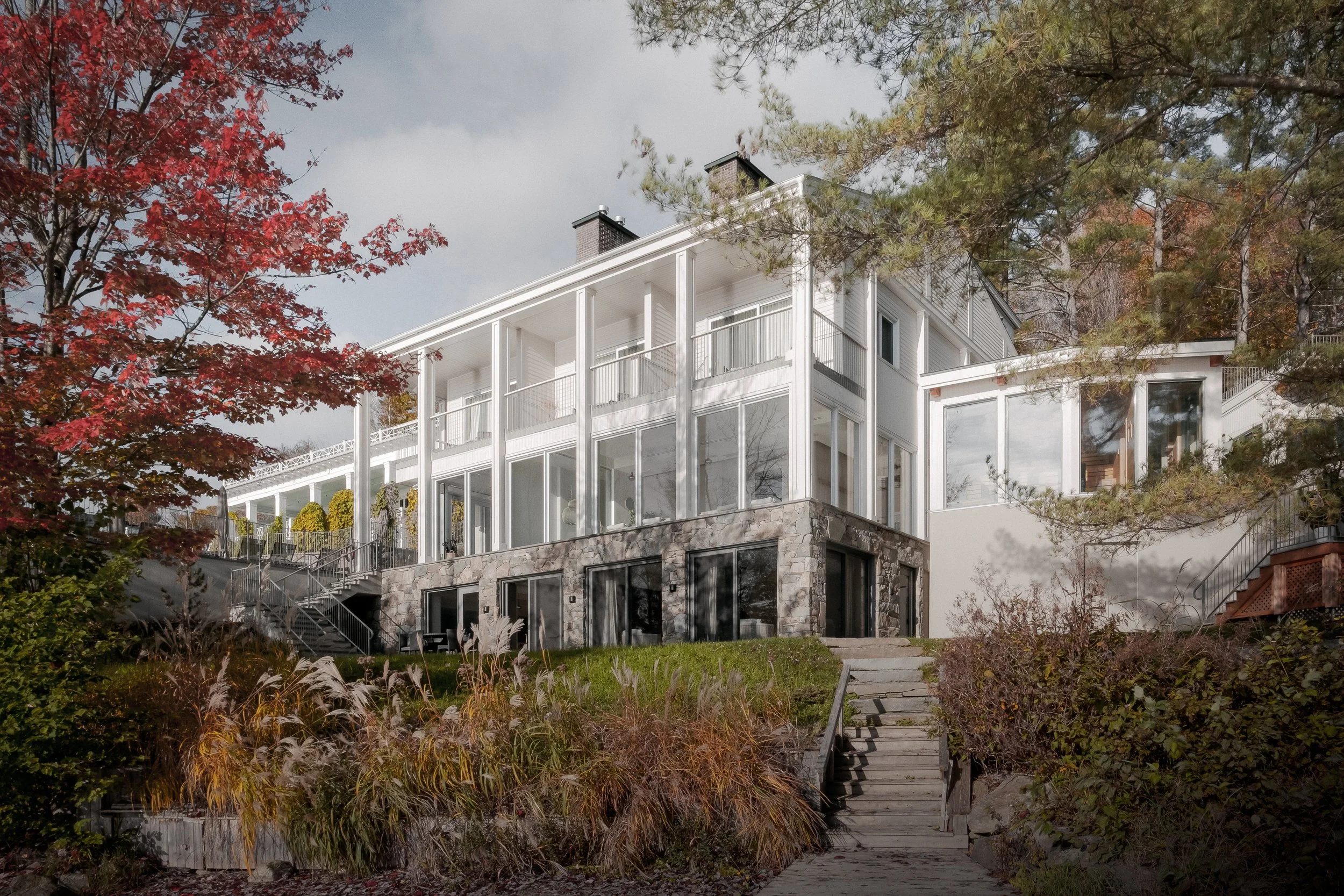 Modern multi-story house with a stone and white exterior, large windows, and a backyard with stairs leading to a garden, surrounded by trees with autumn foliage.