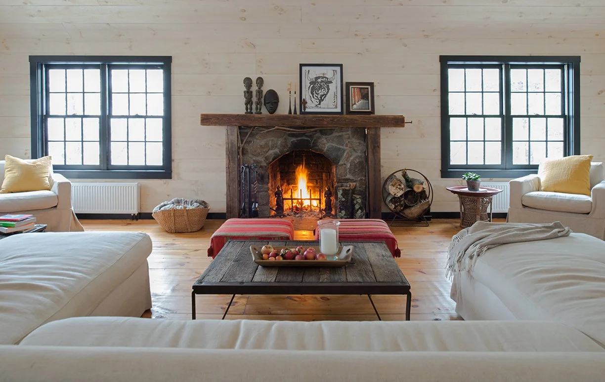 Living room with a lit stone fireplace, two windows, white sofas, a wooden coffee table with apples and a candle, decorative items on the mantel, and a cozy, rustic aesthetic.