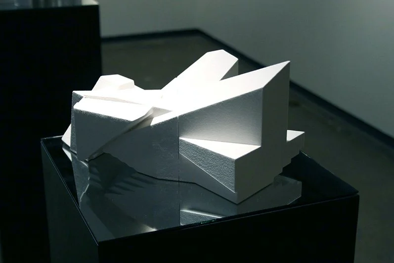 White abstract sculpture with sharp geometric shapes displayed on a black pedestal in a gallery.