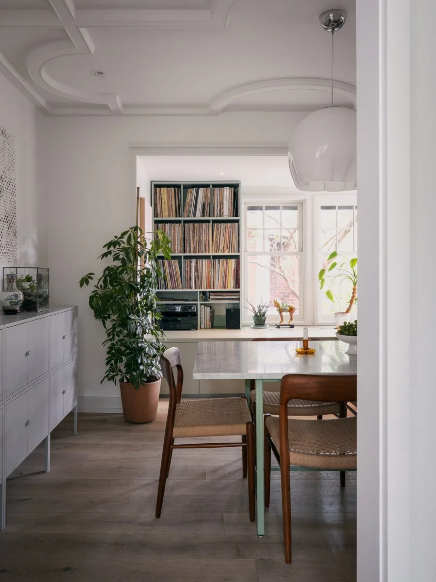 A cozy dining room with a white marble table, wooden chairs, a tall bookshelf filled with vinyl records, and large windows allowing natural light, decorated with potted plants and ornaments.