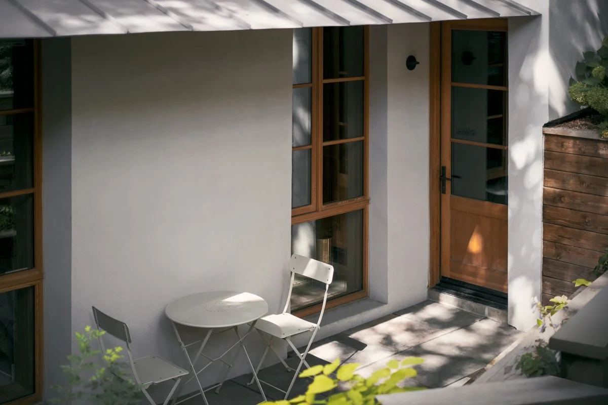 A small outdoor patio area with a white round table and two white folding chairs positioned in front of a house with white walls, large windows with wooden frames, and a wooden door. Shadows of surrounding trees cast across the scene.