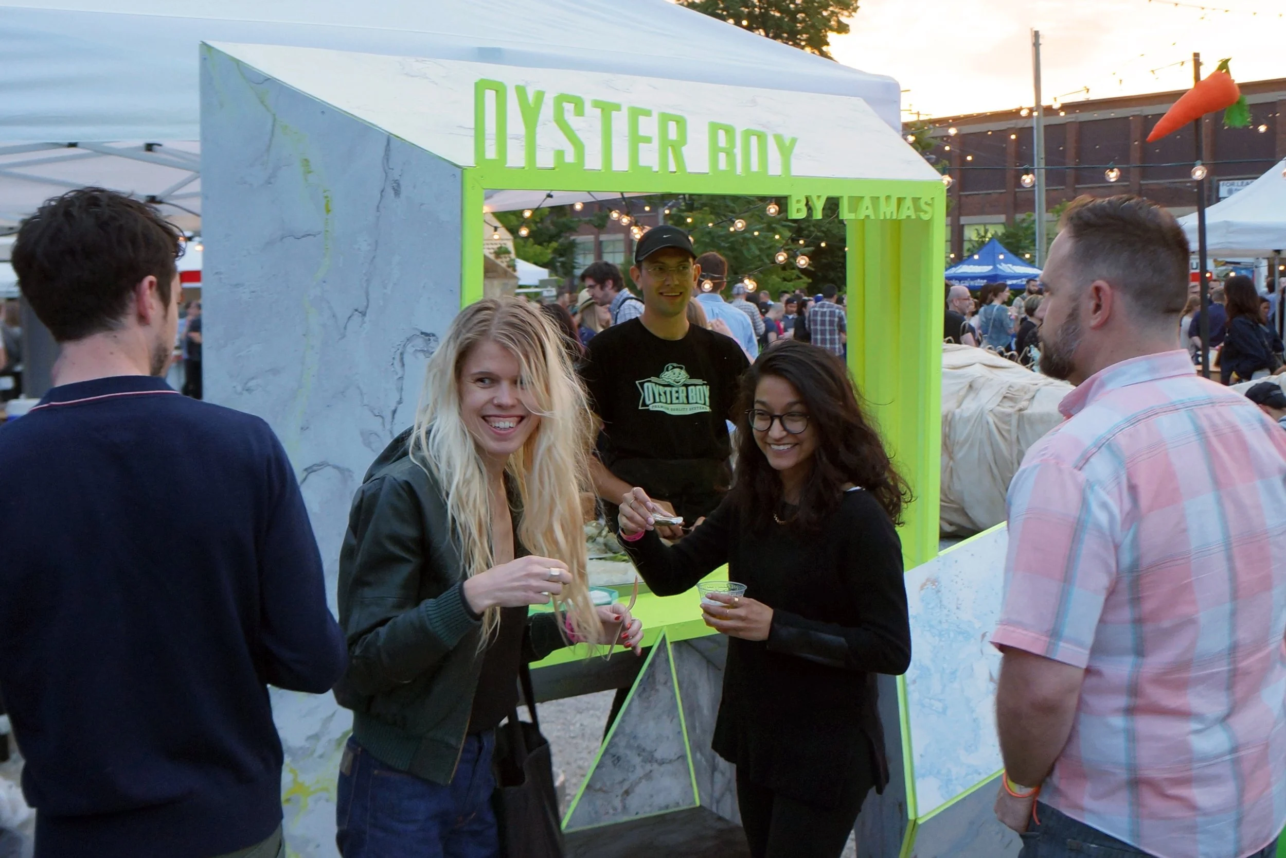 People at a food festival around a booth that says "Oyster Ray by Lamass" selling oysters, with string lights and tents in the background.
