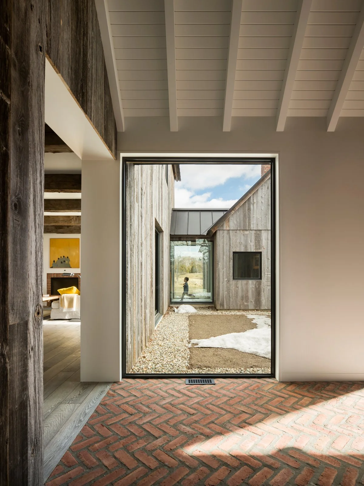 View through a glass door showing a modern outdoor area with gravel, dirt patches, and snow remnants, surrounded by weathered wooden walls of a contemporary house with white interior walls and exposed wooden beams ceiling.