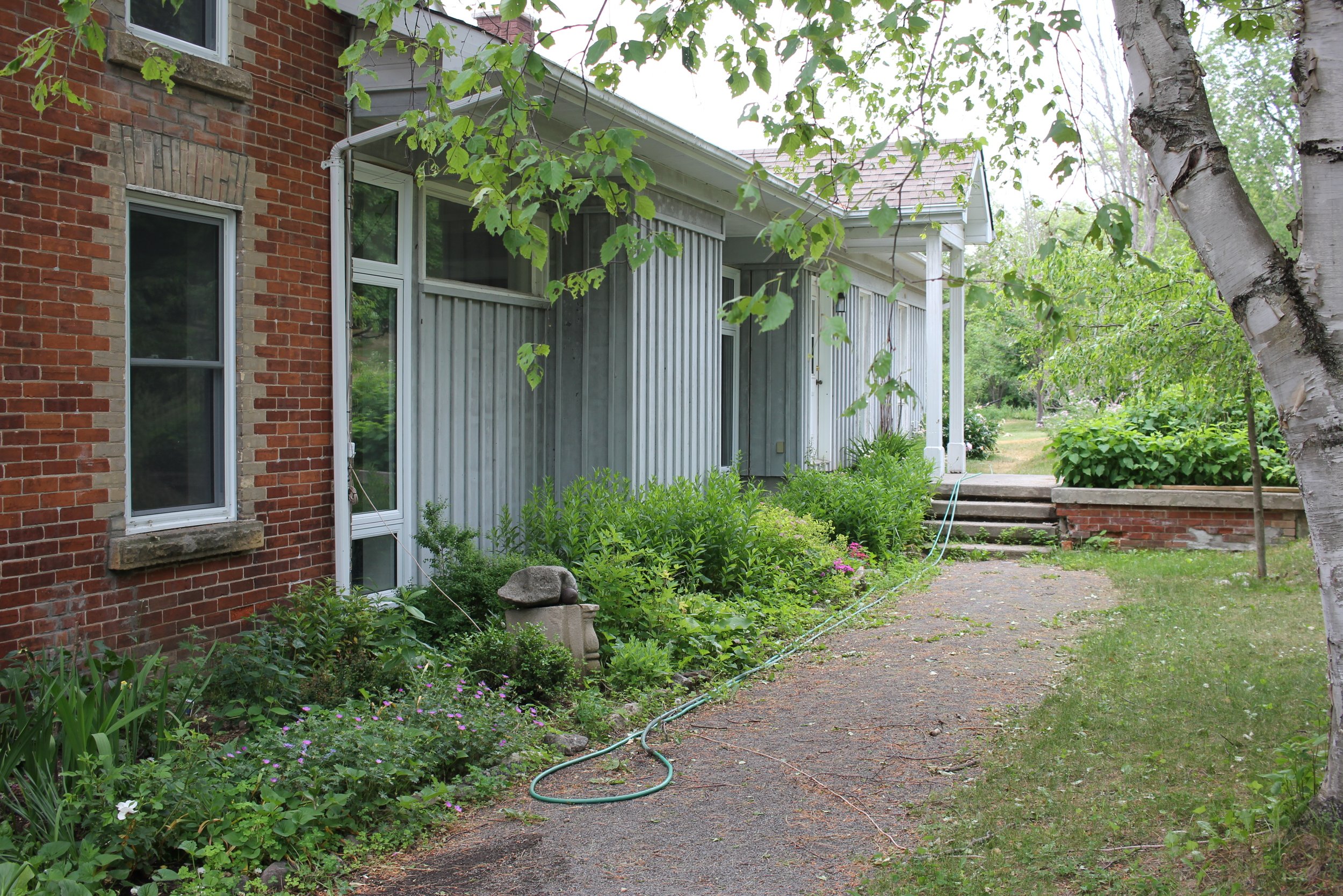 A residential backyard with a garden, brick and gray wooden siding house, trees, and a gravel pathway.