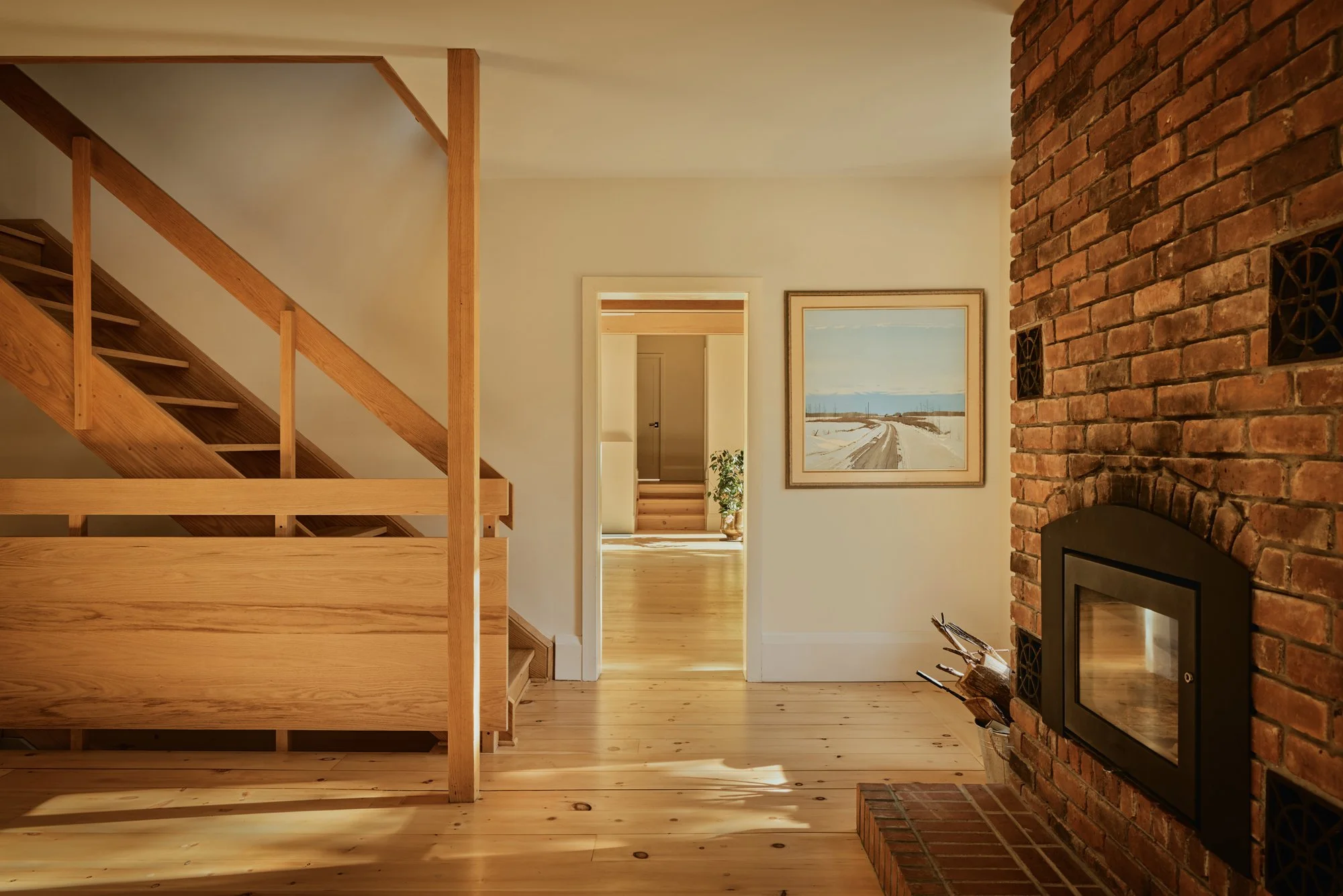 Interior view of a cozy living room with a brick fireplace, wooden staircase, wooden floors, and artwork on the wall, with sunlight streaming in.
