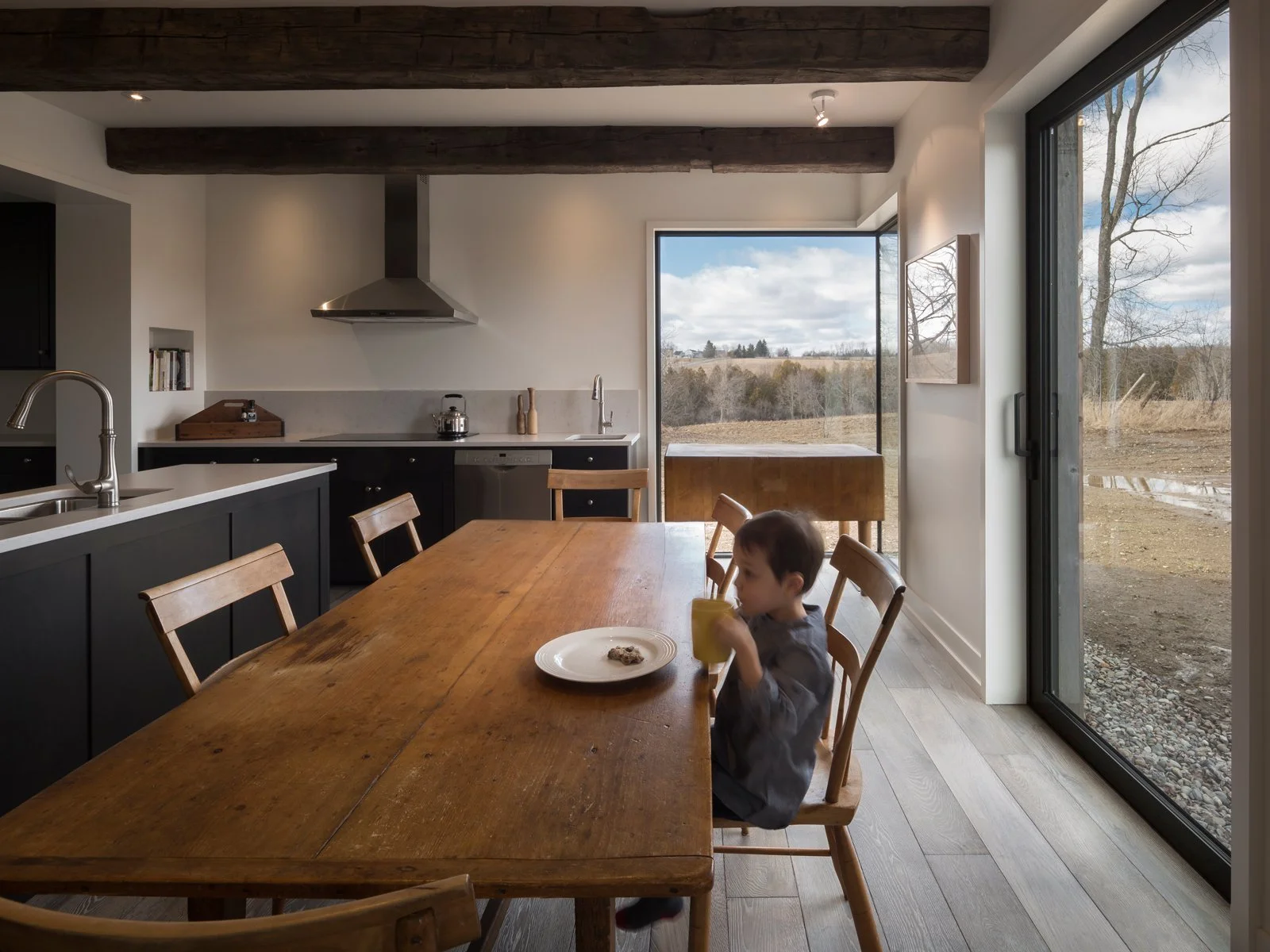 A young boy sitting at a wooden dining table drinking from a yellow cup with a plate of cookies in front of him, inside a modern kitchen with large windows showing an outdoor view of trees and fields.
