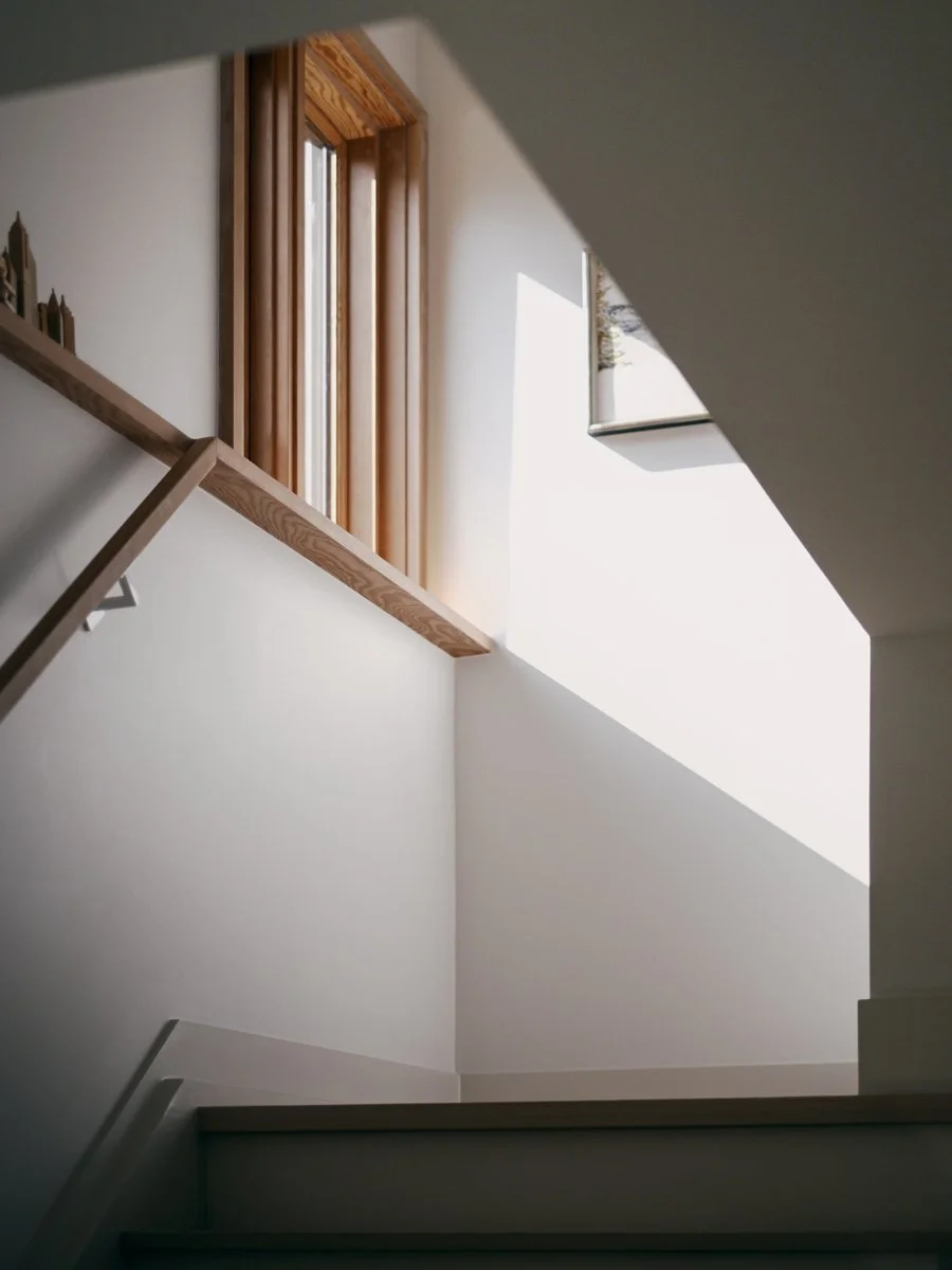 Interior view of a staircase with a large wooden-framed window allowing natural light, white walls, and a wall-mounted art piece visible.