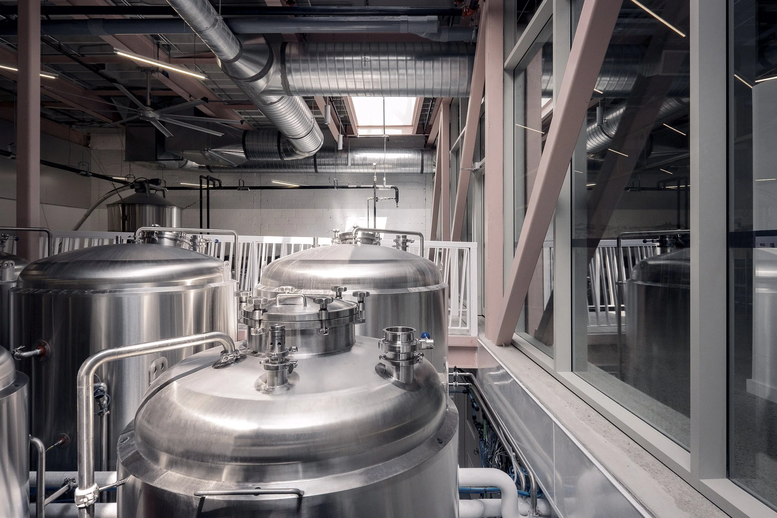 Interior view of a brewery with large stainless steel fermentation tanks and ductwork visible on the ceiling.