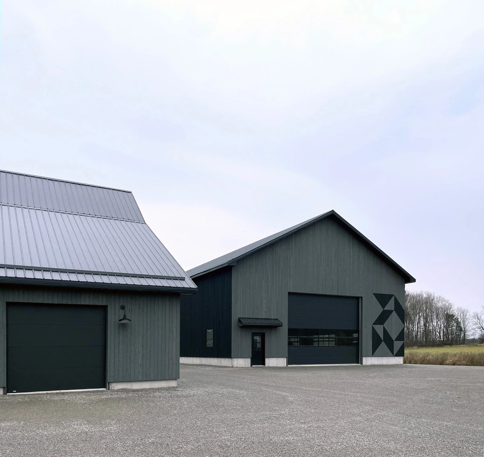 Two modern buildings with dark gray exteriors and metal roofs, one with a geometric mural on the wall, situated on a gravel lot with open fields in the background.
