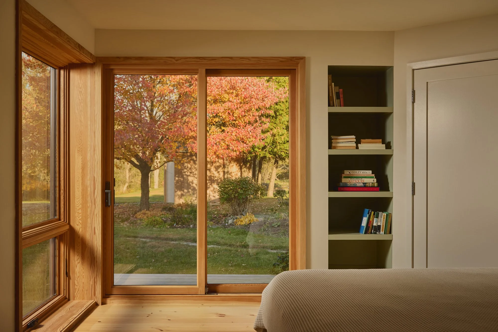 Interior of a room with large sliding glass door and window showing a tree with colorful fall foliage outside, wooden trim, and a built-in bookshelf filled with books.