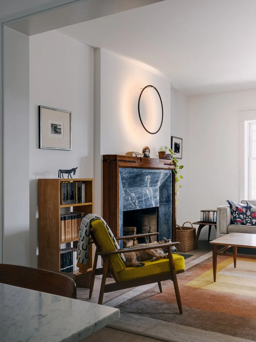 Living room with a mid-century modern style. Features a yellow armchair with a dog lying on it, a wooden coffee table, a blue marble fireplace with a wooden mantel, a bookshelf, and wall art. Natural light from a nearby window illuminates the space.