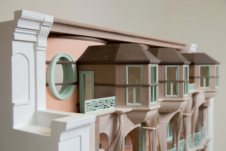 Architectural model of a building with detailed facades and bay windows, displayed on a table with sketchbooks in the background.