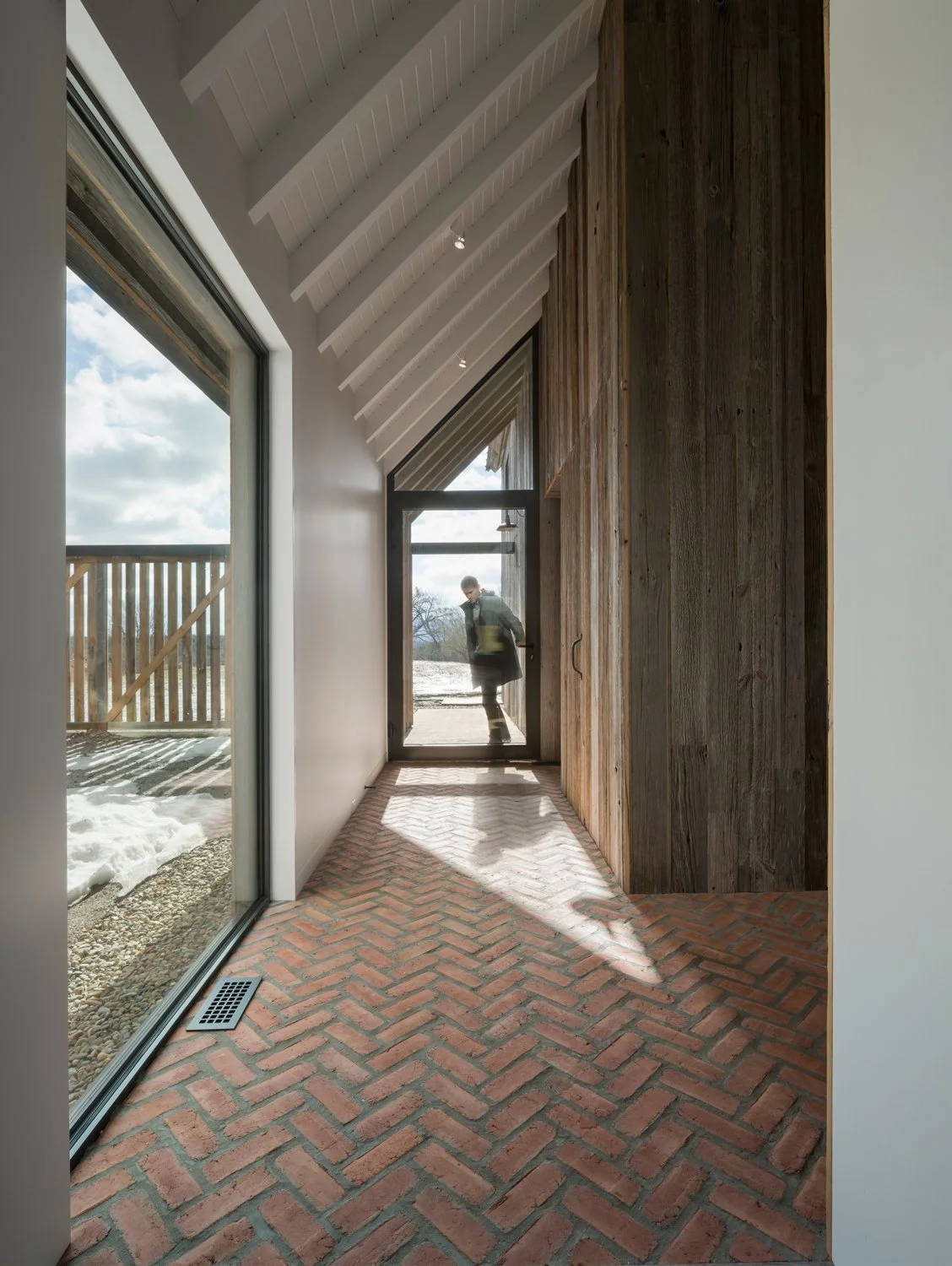 Interior view of a modern hallway with brick floor, large window on the left, wall with wooden paneling on the right, and glass door at the end with a person standing outside.