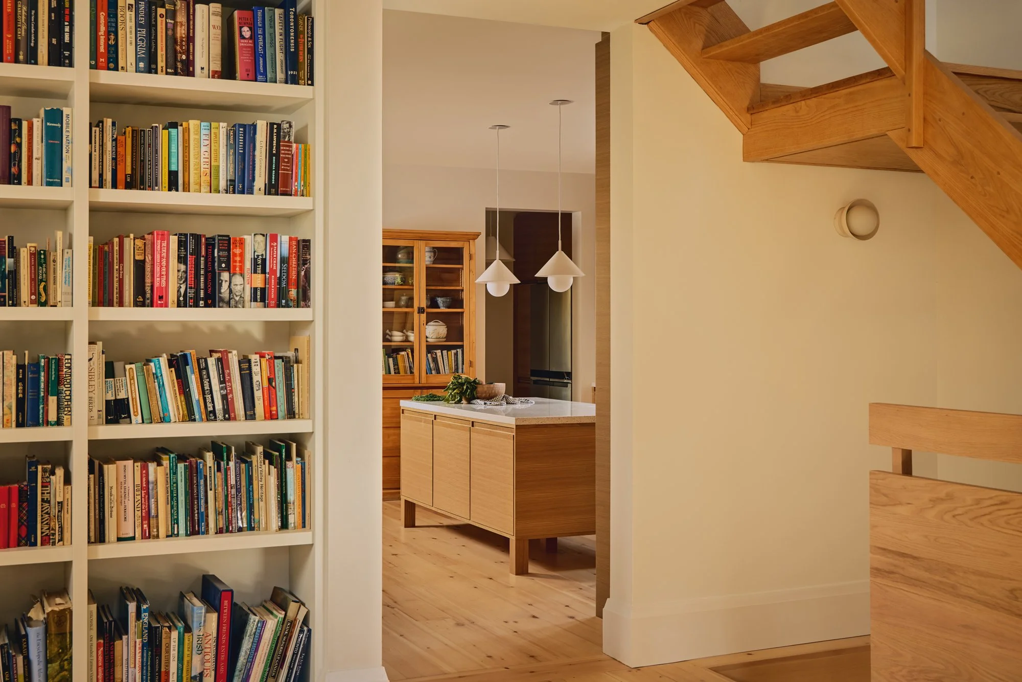 Interior view of a home with a built-in white bookshelf filled with colorful books on the left, a hallway leading to a kitchen with a wooden island and cabinets, pendant lights hanging above, and a staircase with wooden steps on the right.
