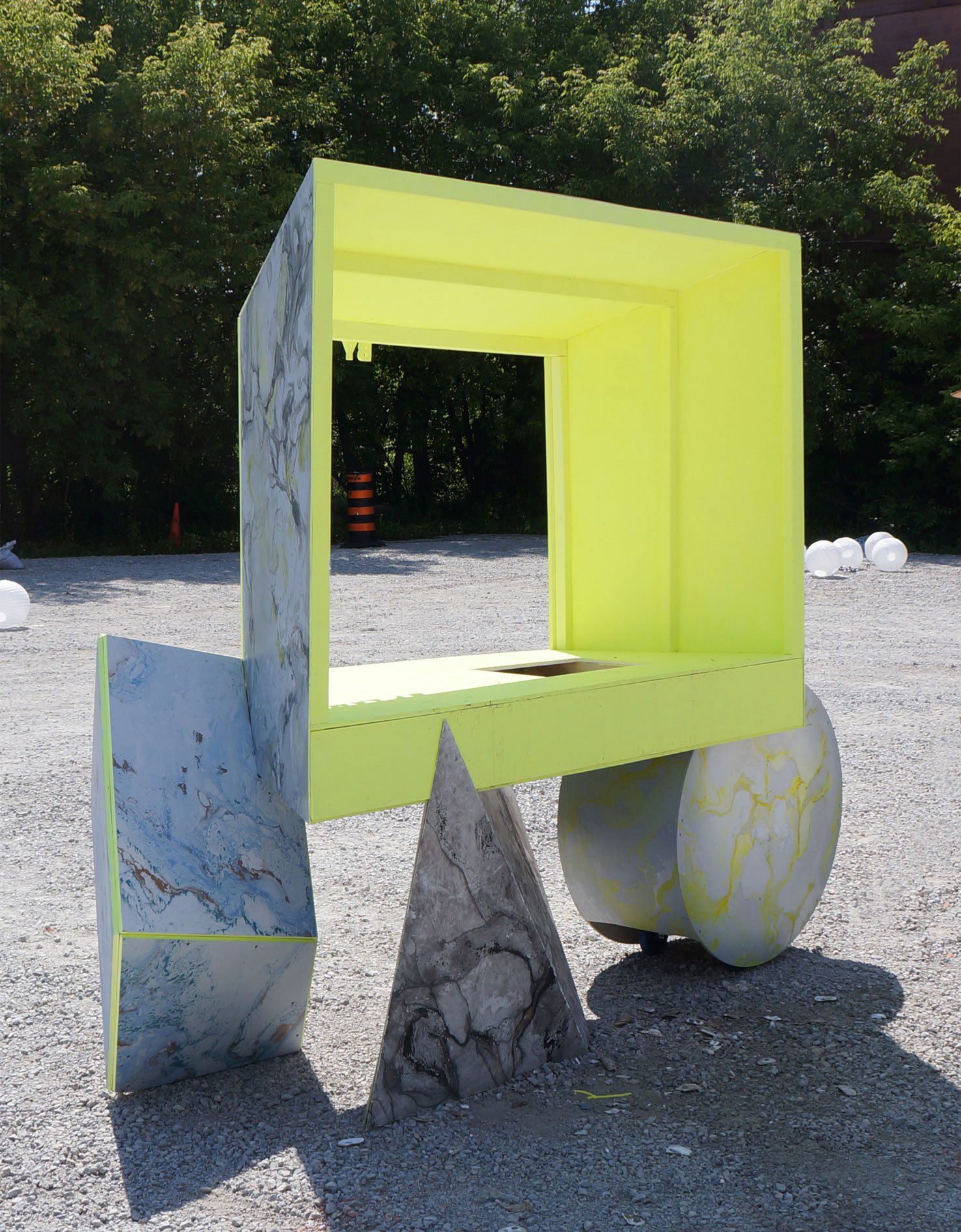 An outdoor art installation featuring a large, colorful geometric structure with a square yellow frame, marble-patterned panels, and variously shaped support elements on a gravel surface.